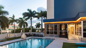 Una piscina con una silla de salvavidas, un área de estar cubierta y un edificio con ventanas amplias y una pared azul decorativa.