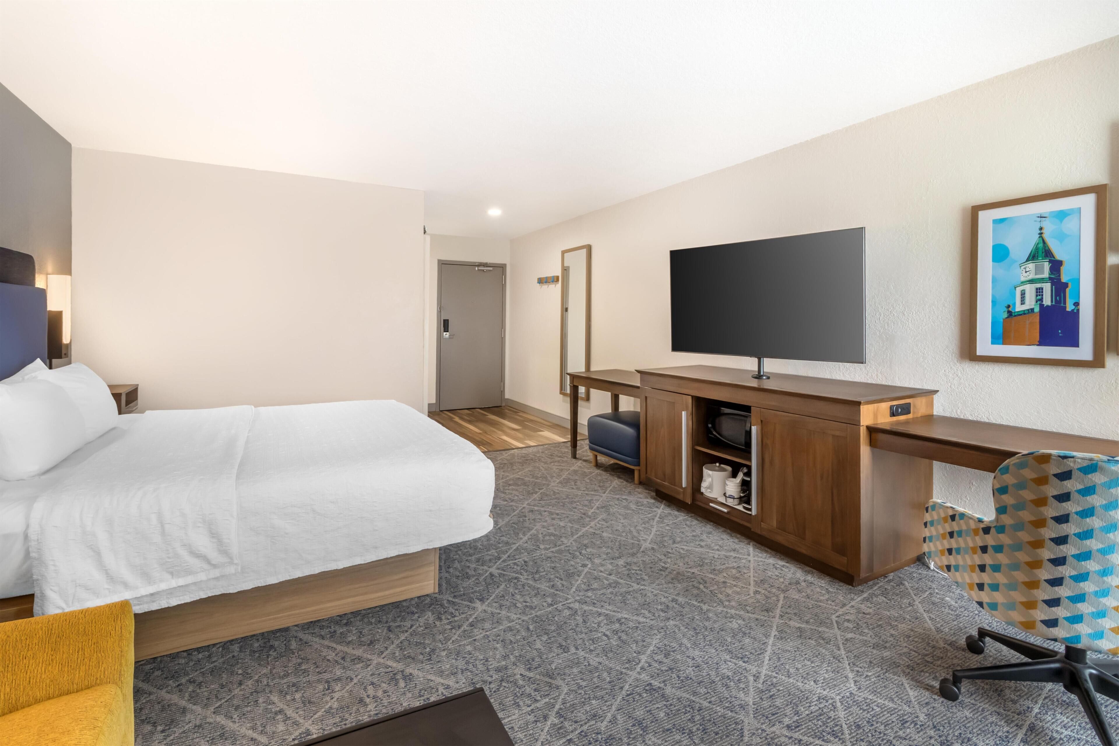 A hotel room with a bed, a desk, a chair, a television, and a painting on the wall.