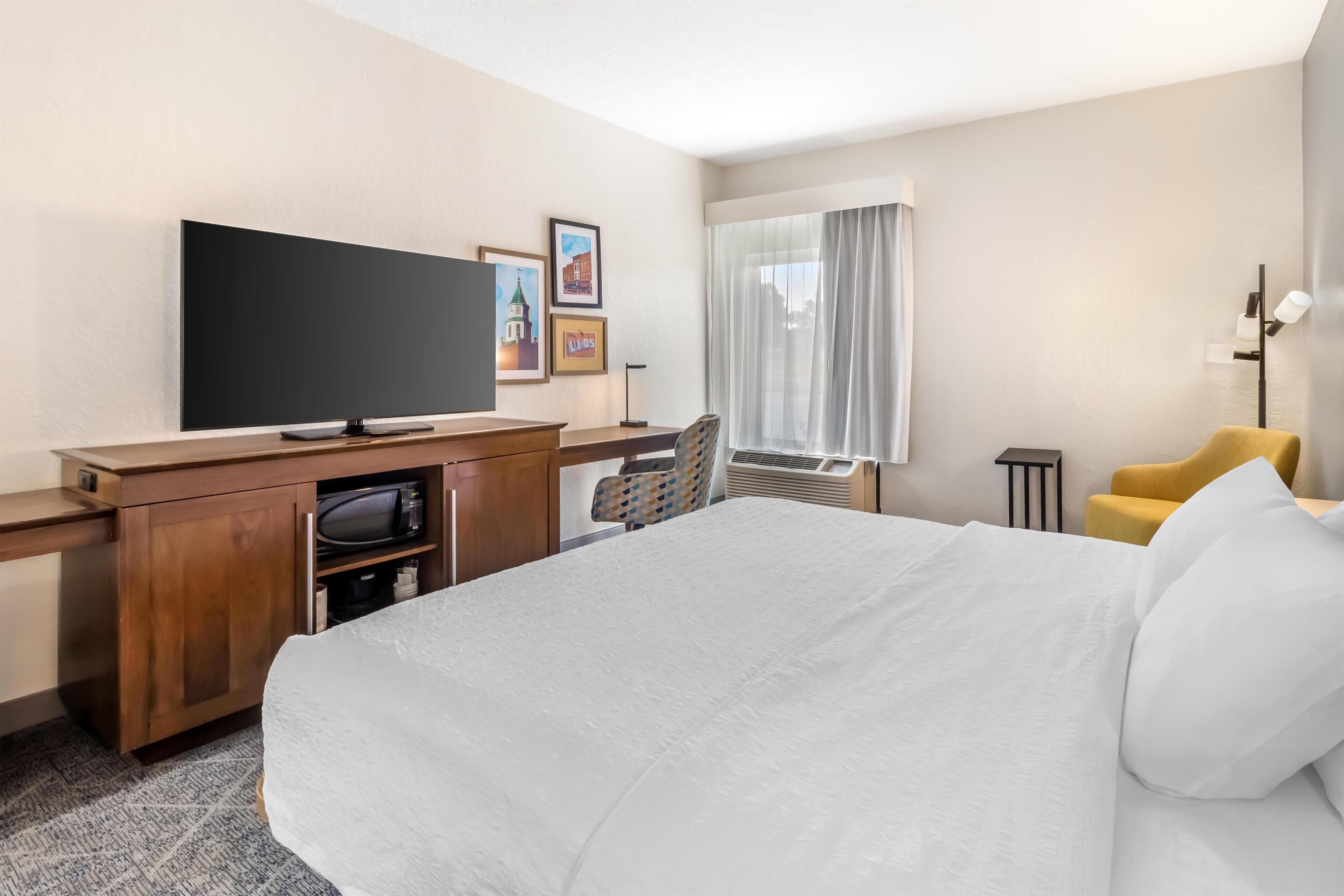 A hotel room with a bed, a desk with a flat-screen TV, a chair, a window with curtains, and a wall-mounted lamp.