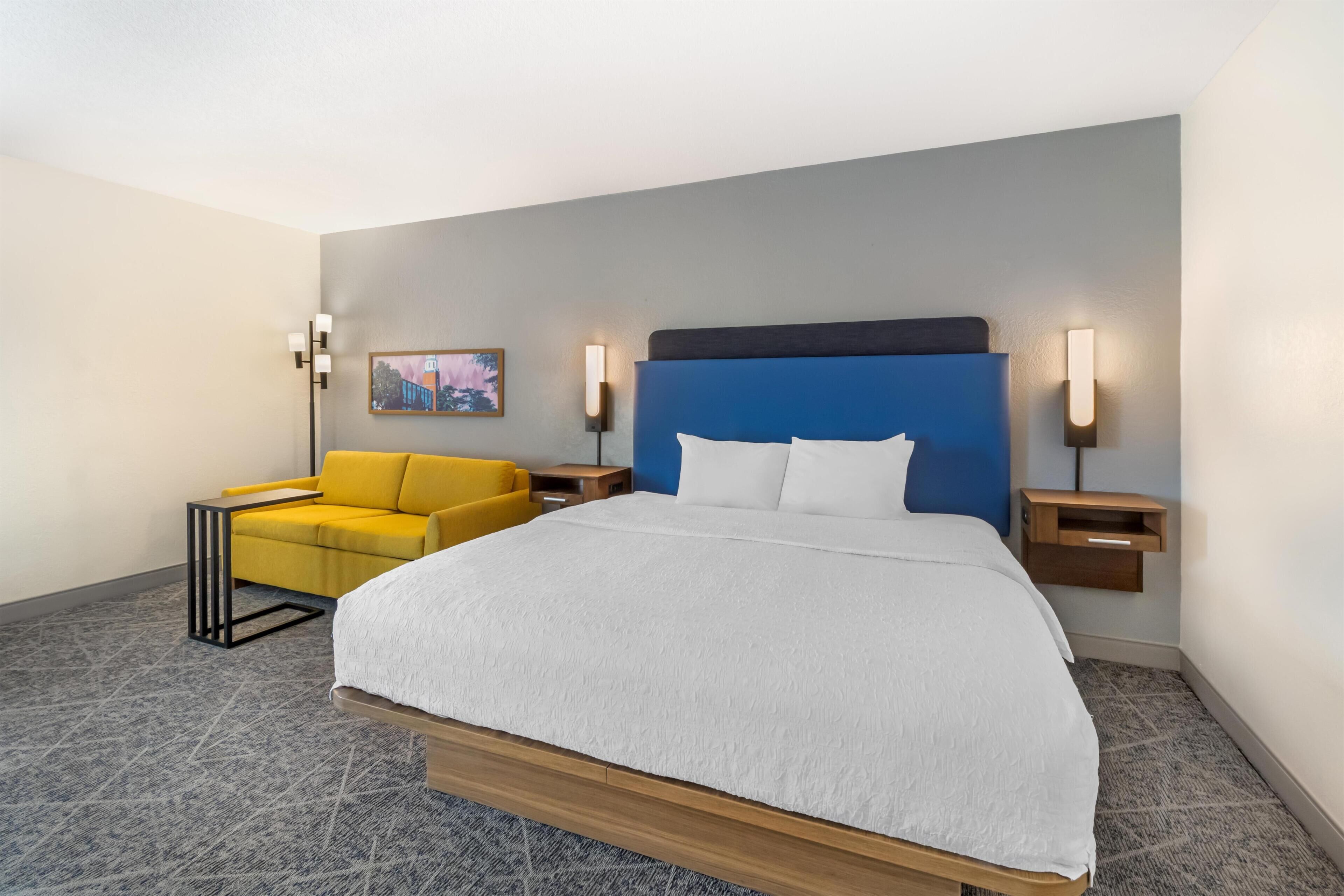 Quarto de hotel com uma cama grande, uma poltrona amarela e duas mesas de cabeceira com luminárias fixadas na parede.