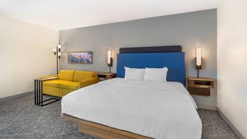 Quarto de hotel com uma cama grande, uma poltrona amarela e duas mesas de cabeceira com luminárias fixadas na parede.