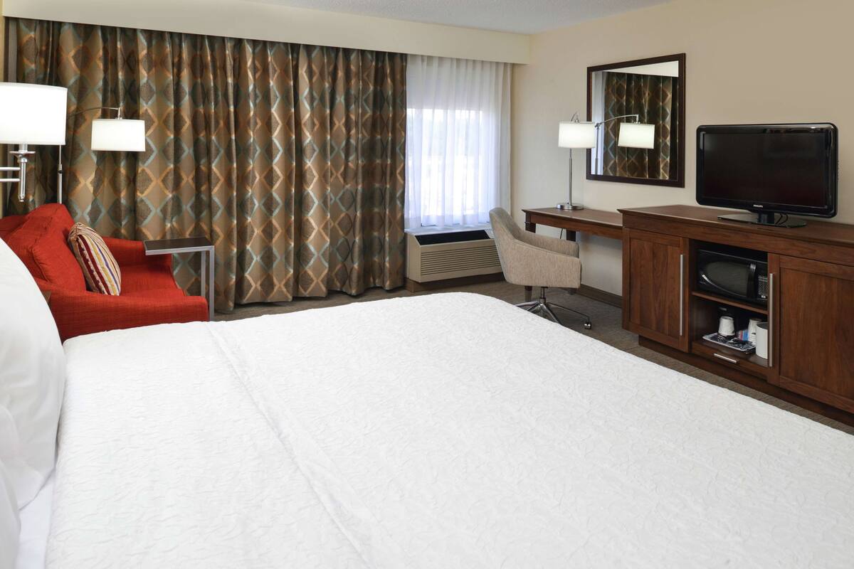 A hotel room with a large bed, a red armchair, a desk with a chair, a television, and a large window with curtains.