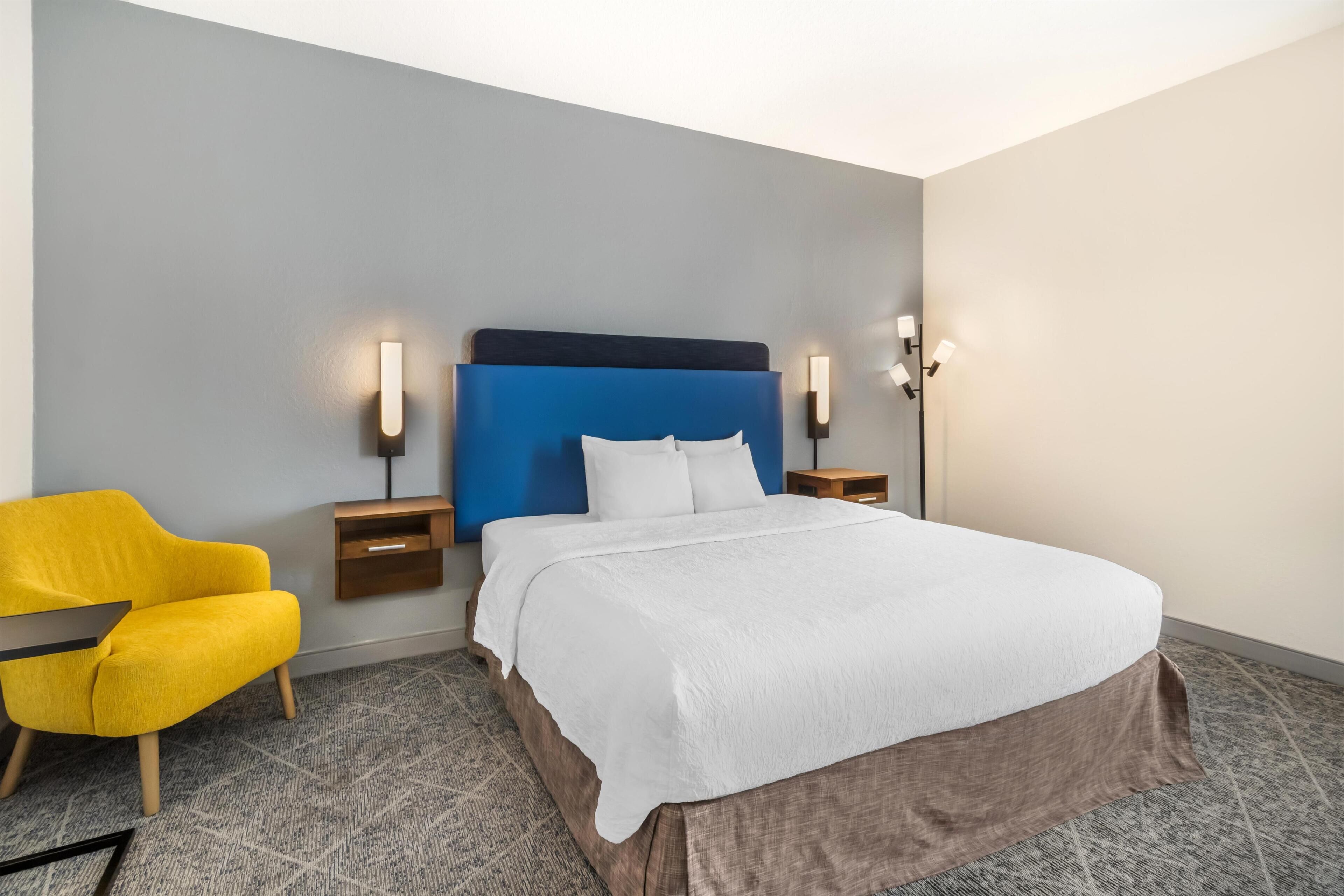 A hotel room with a bed, a nightstand, a lamp, a chair, and a carpeted floor.
