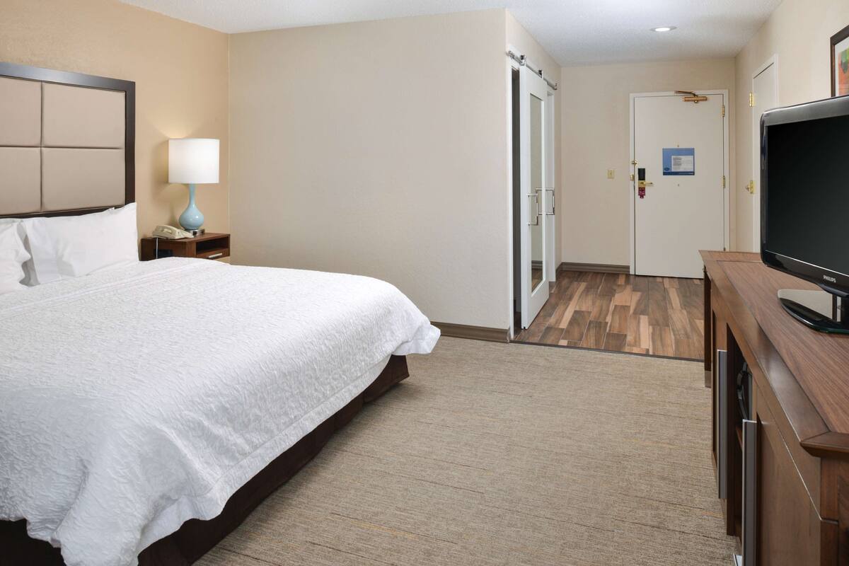 A hotel room with a large bed, a bedside table with a lamp, a television, and a door leading to another room.