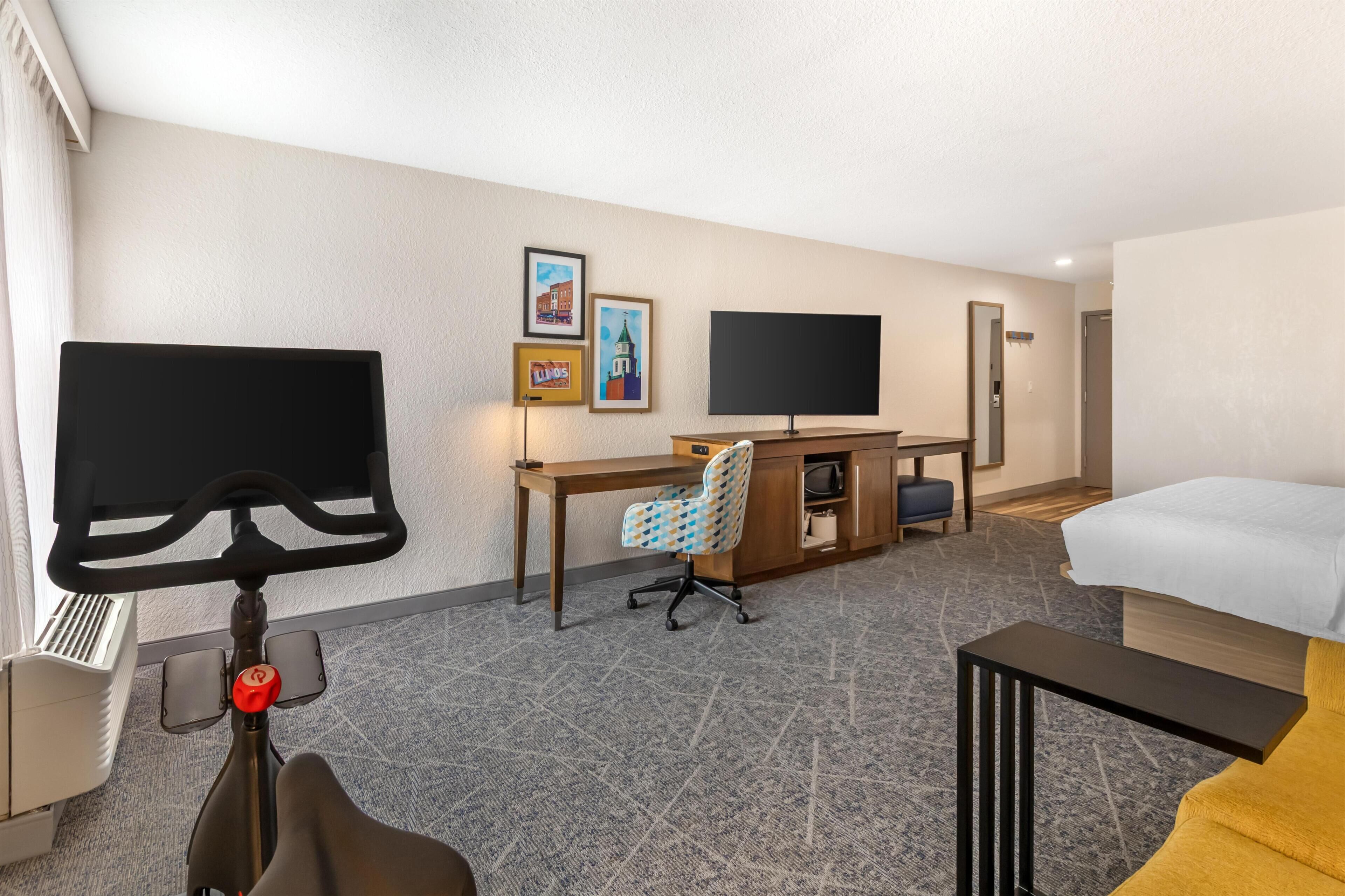 Habitación de hotel con cama, escritorio, silla, televisión y televisor montado en la pared.