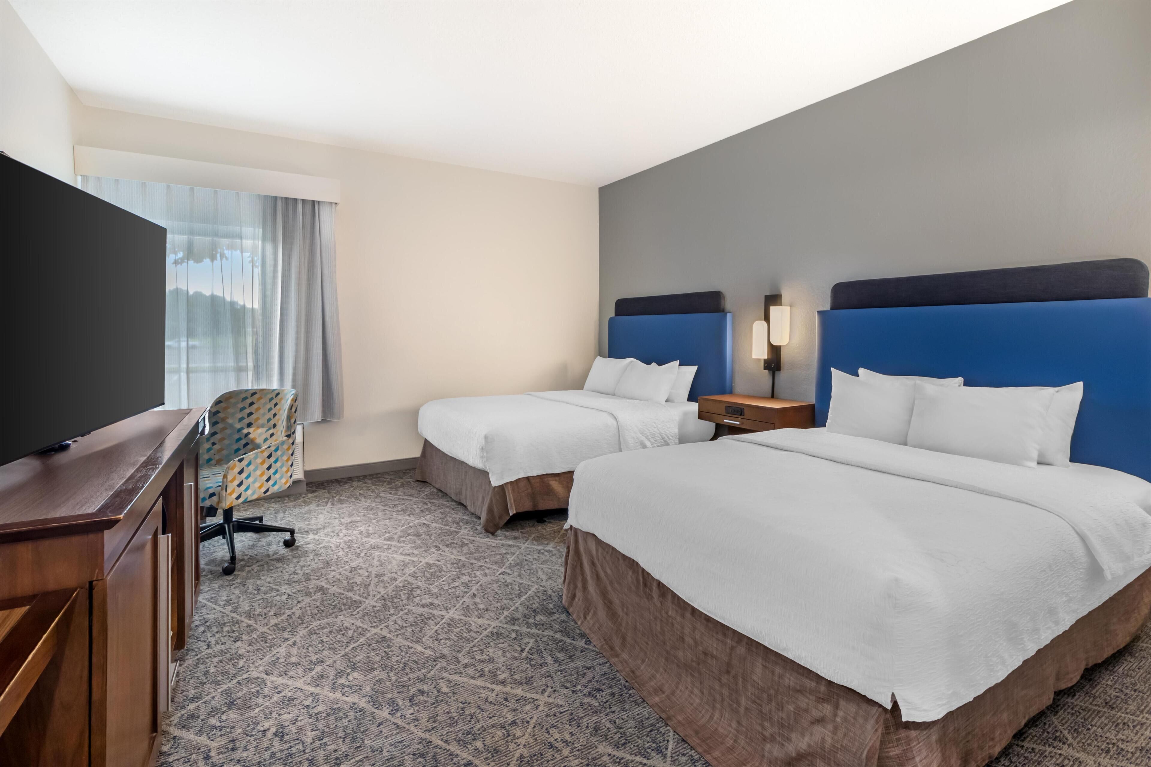 A hotel room with two beds, a desk, a chair, and a TV.