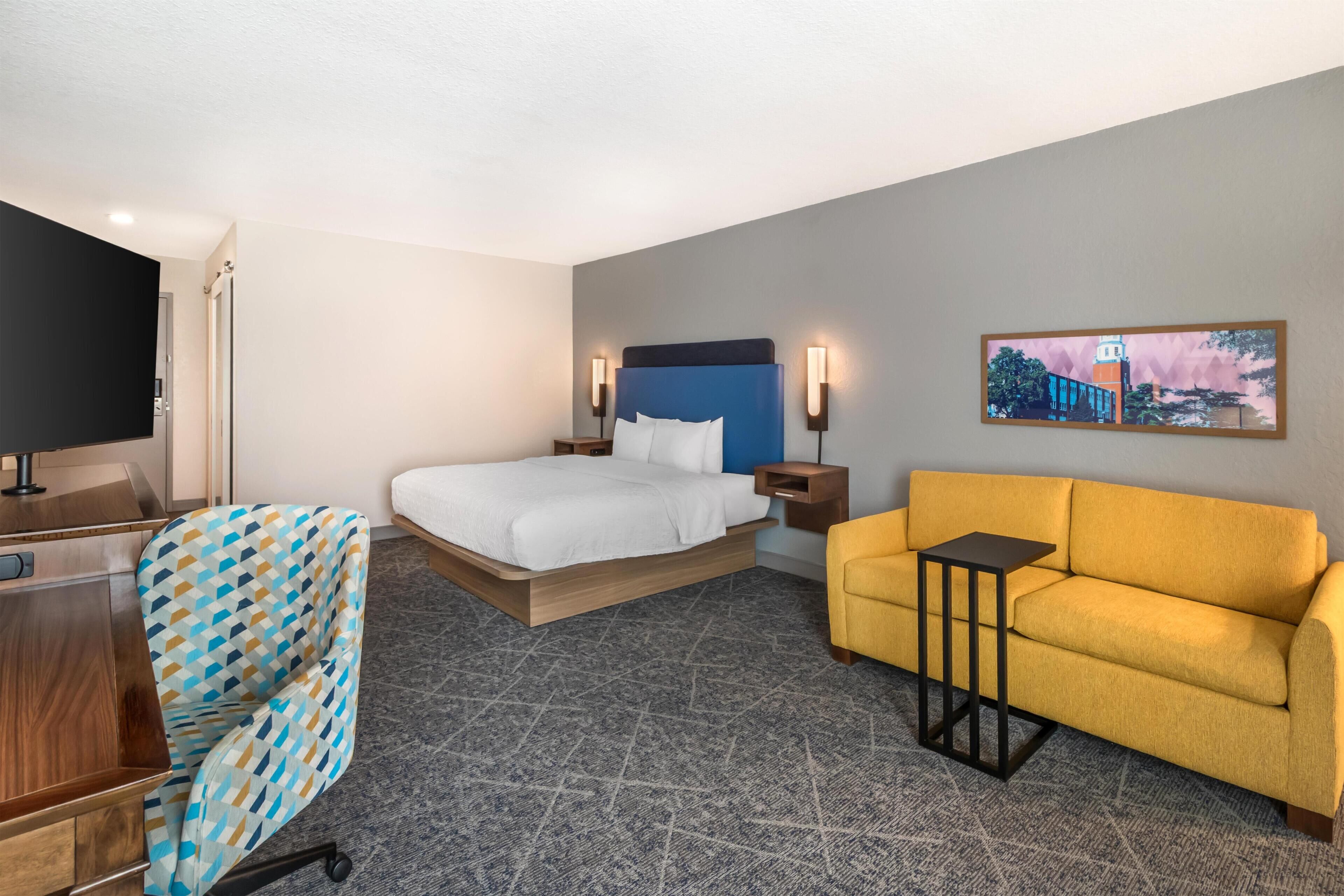 A hotel room with a bed, a sofa, a desk, a chair, and a TV.