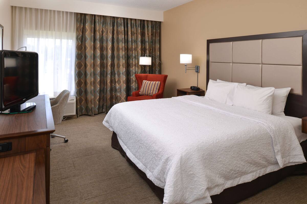 A hotel room with a large bed, a TV on a desk, a chair, and a nightstand with a lamp.
