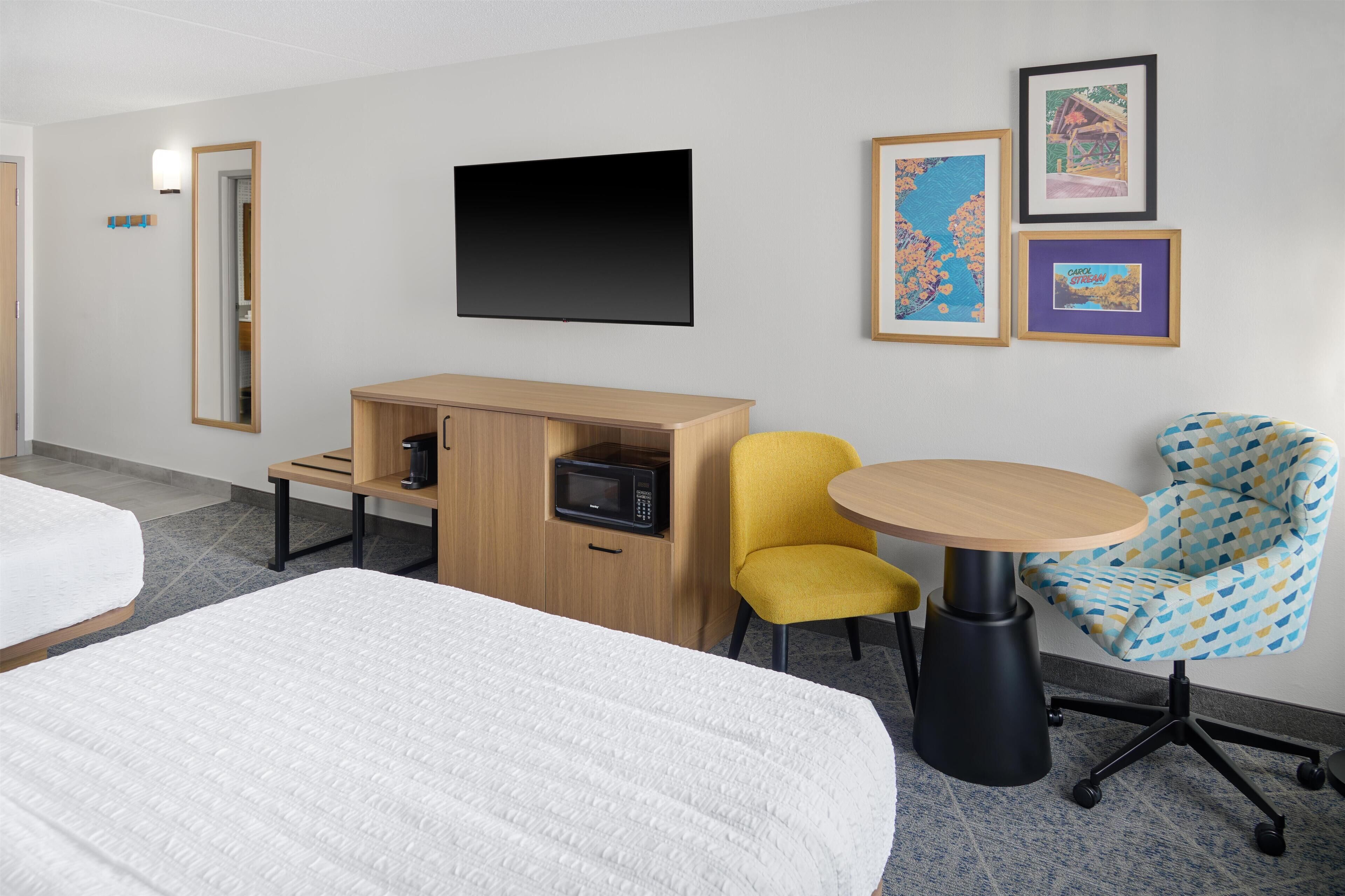 Habitación de hotel con dos camas, un escritorio con microondas, una silla amarilla, una mesa redonda y cuadros en la pared.