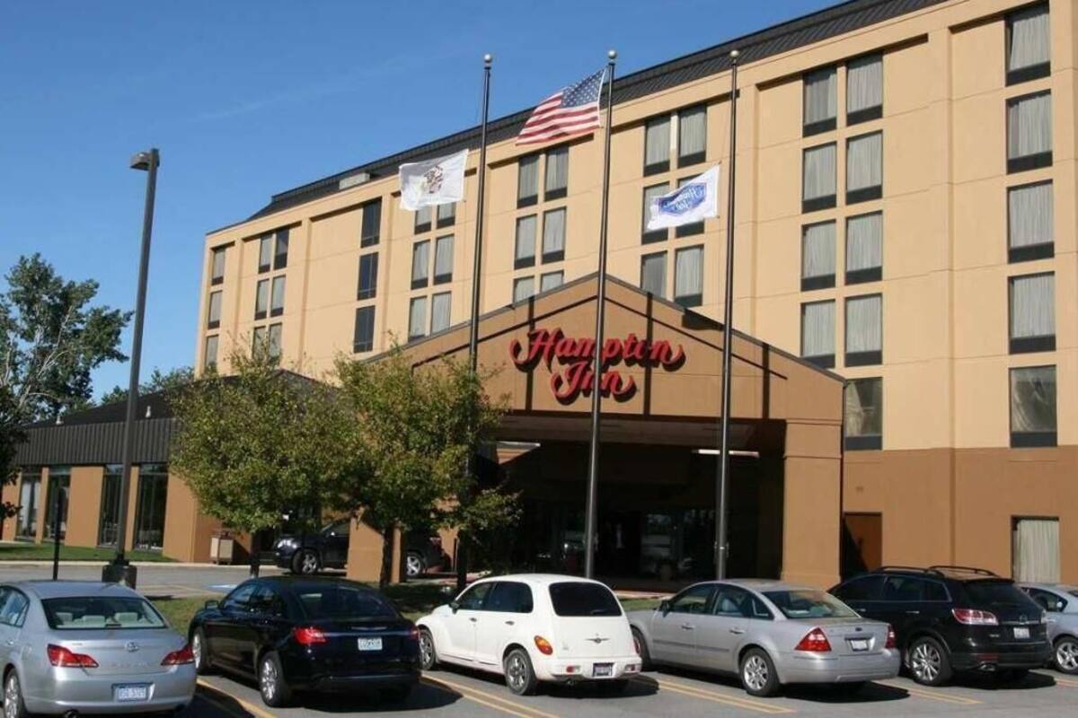 Um hotel Hampton Inn com entrada acessível, bandeiras americanas e um estacionamento com vários carros.