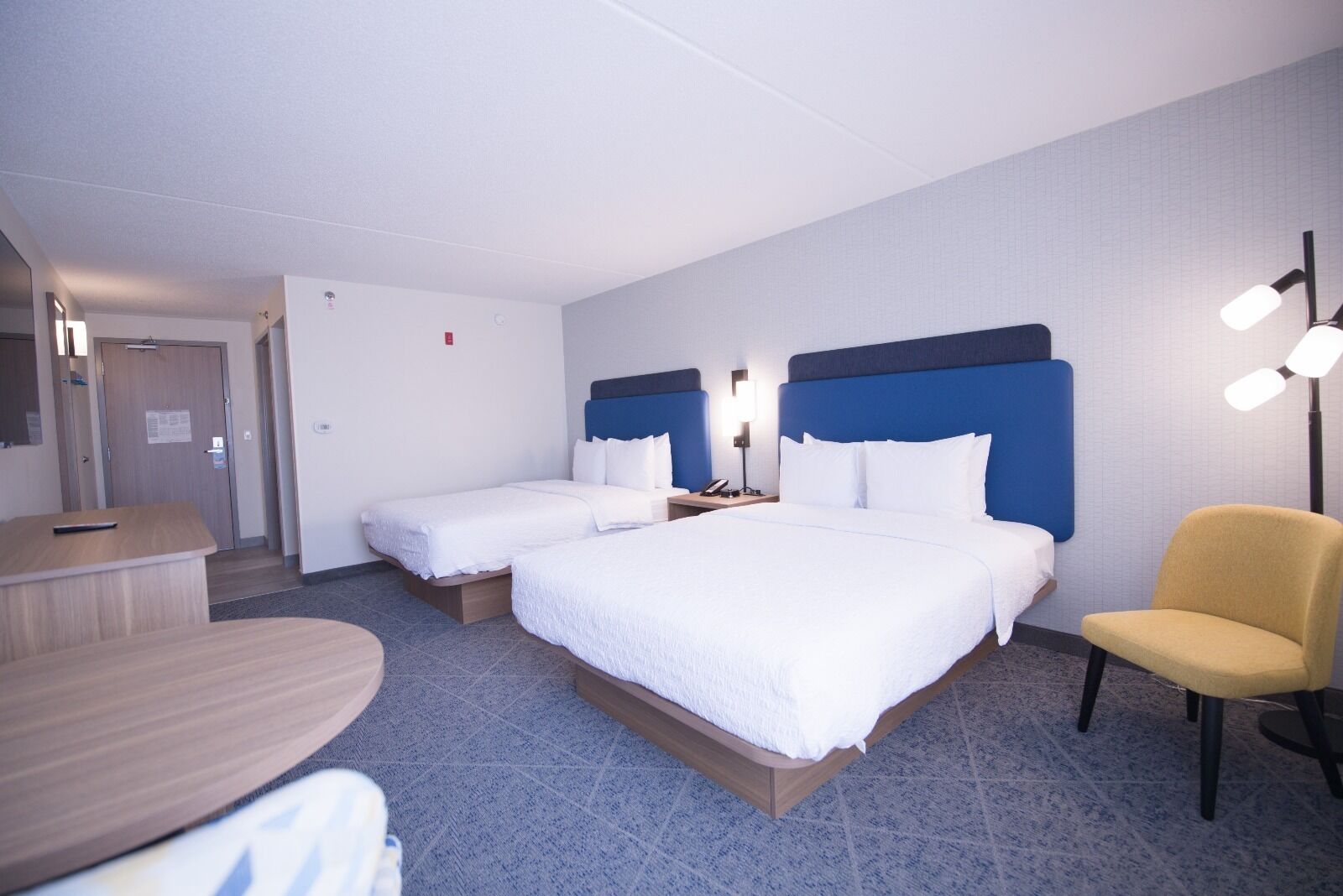 A hotel room with two beds, a desk, a chair, and a nightstand with a lamp.