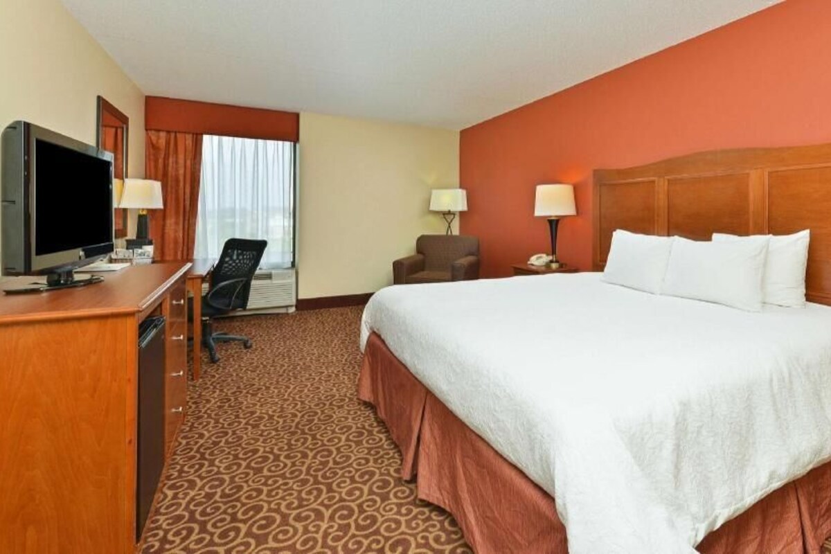 A hotel room with a bed, a desk with a television, a chair, and a lamp.