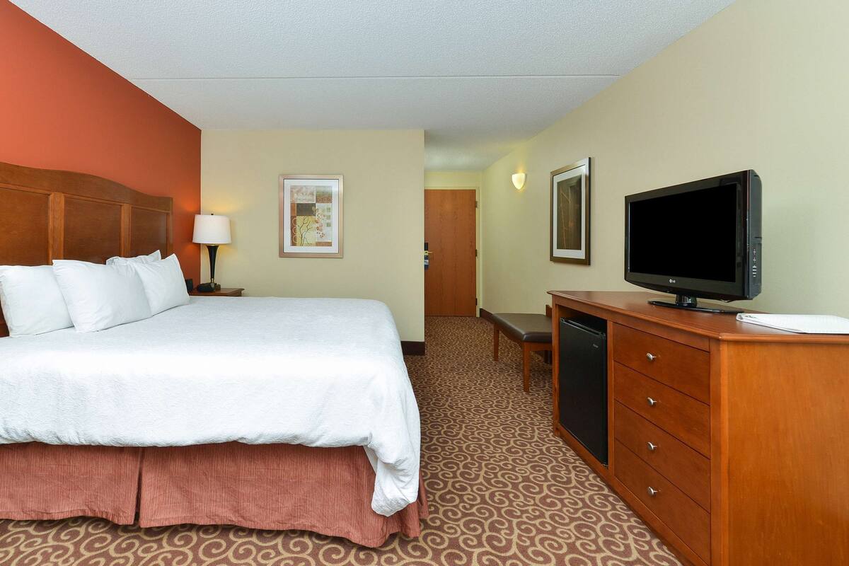 Quarto de hotel com uma cama, uma mesa de trabalho com televisão, um abajur e uma porta que dá para outro cômodo.