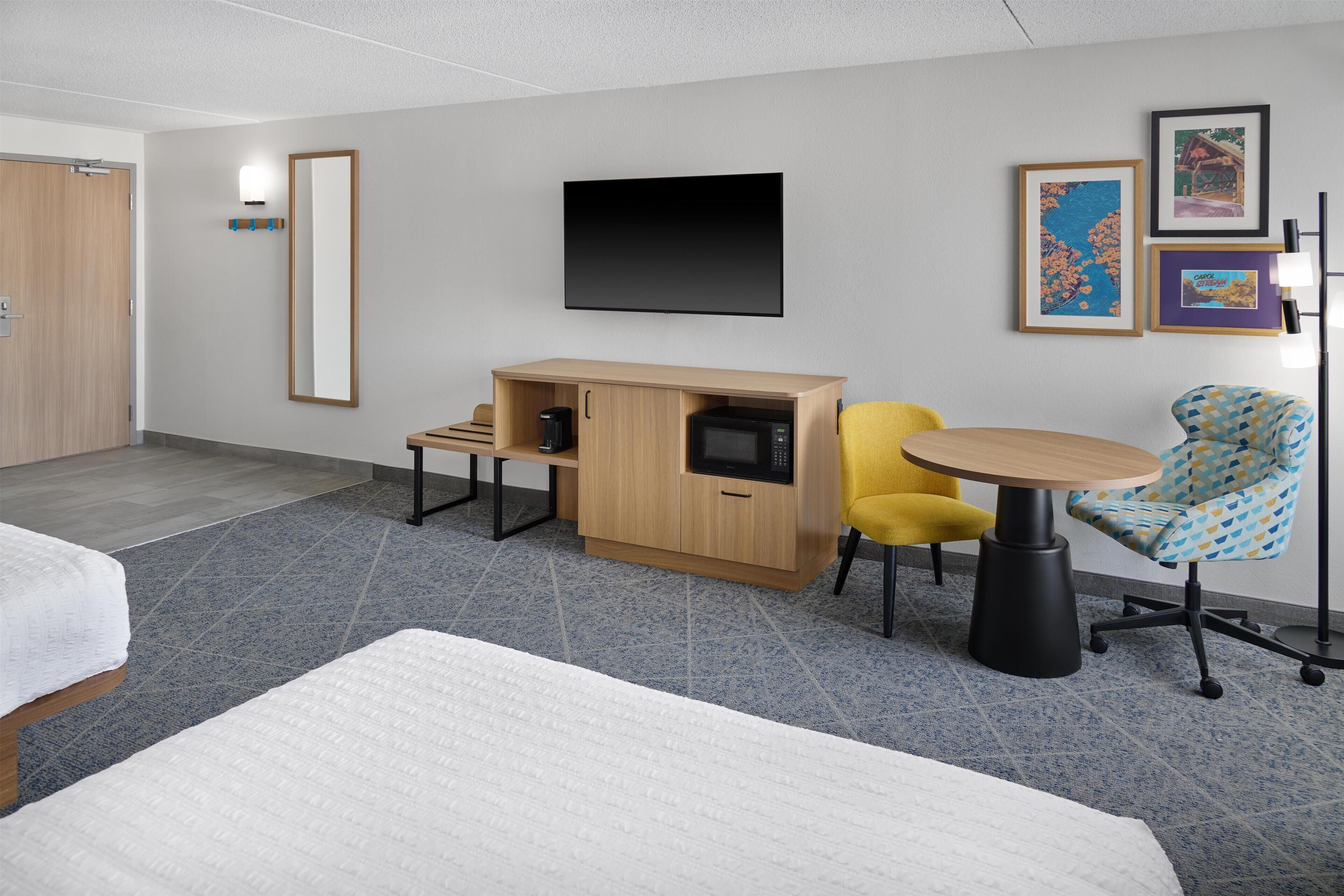A modern hotel room with a bed, a desk with a microwave, a chair, and artwork on the wall.