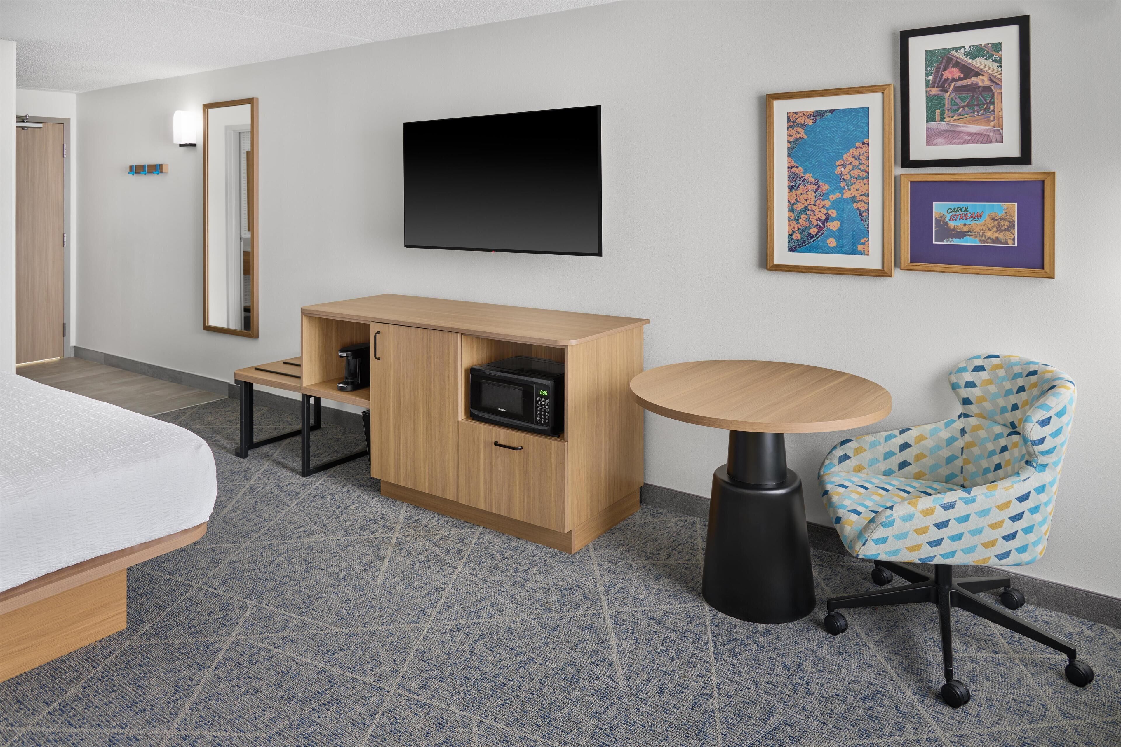 A hotel room with a bed, a wooden desk with a microwave, a round table, a patterned chair, and framed pictures on the wall.