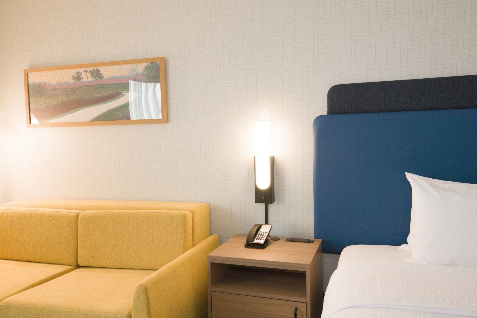 A hotel room with a yellow sofa, a bed with a blue headboard, a bedside table with a lamp and phone, and a painting on the wall.
