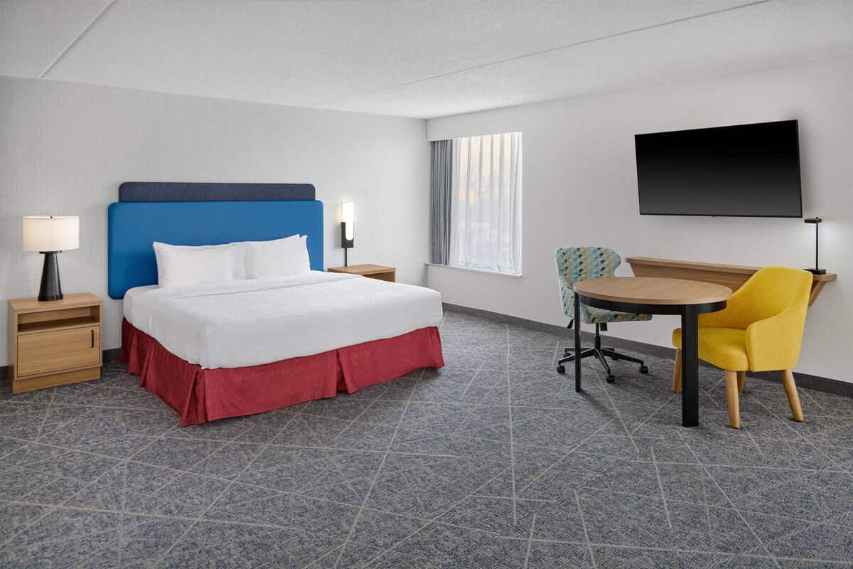 Quarto de hotel com cama, escrivaninha, cadeira, televisão e janela.