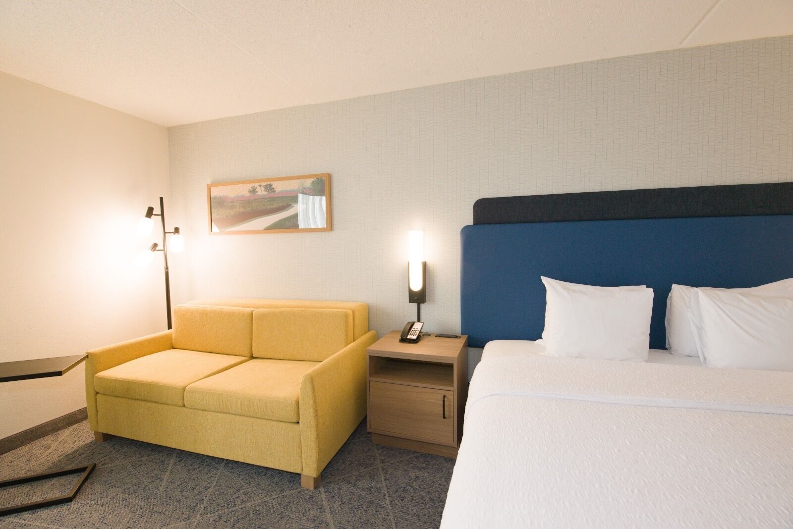 Quarto de hotel com cama, sofá, mesa de cabeceira com abajur e um quadro na parede.