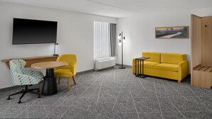 A modern room with a flat-screen TV, a yellow sofa, a round wooden table, a patterned armchair, and a painting on the wall. - Hampton Inn Chicago-Carol Stream (Carol Stream)