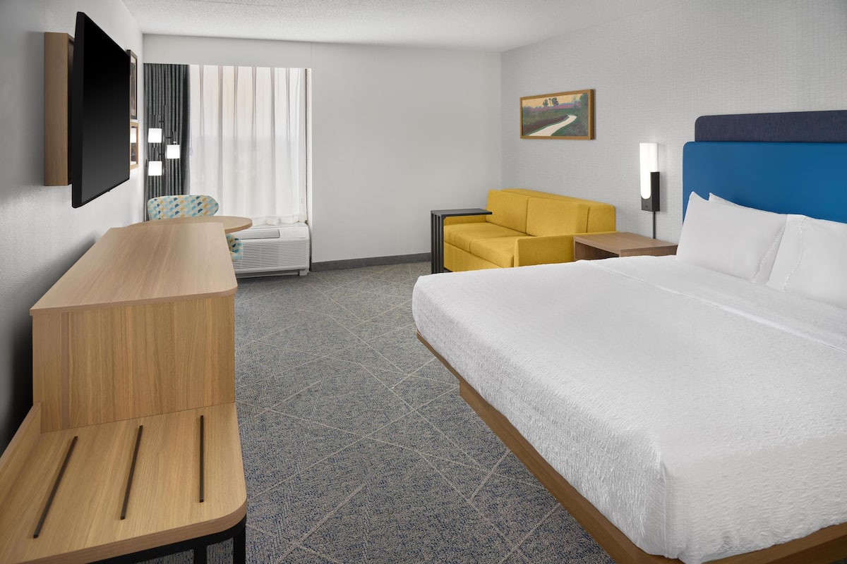 Quarto de hotel com cama grande, escrivaninha, cadeira, televisão e um quadro na parede.