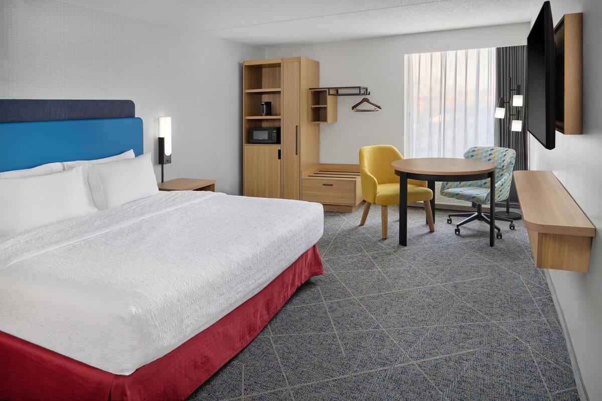 Quarto de hotel com uma cama grande, um guarda-roupa de madeira, uma escrivaninha com cadeira, televisão e uma janela com cortinas.