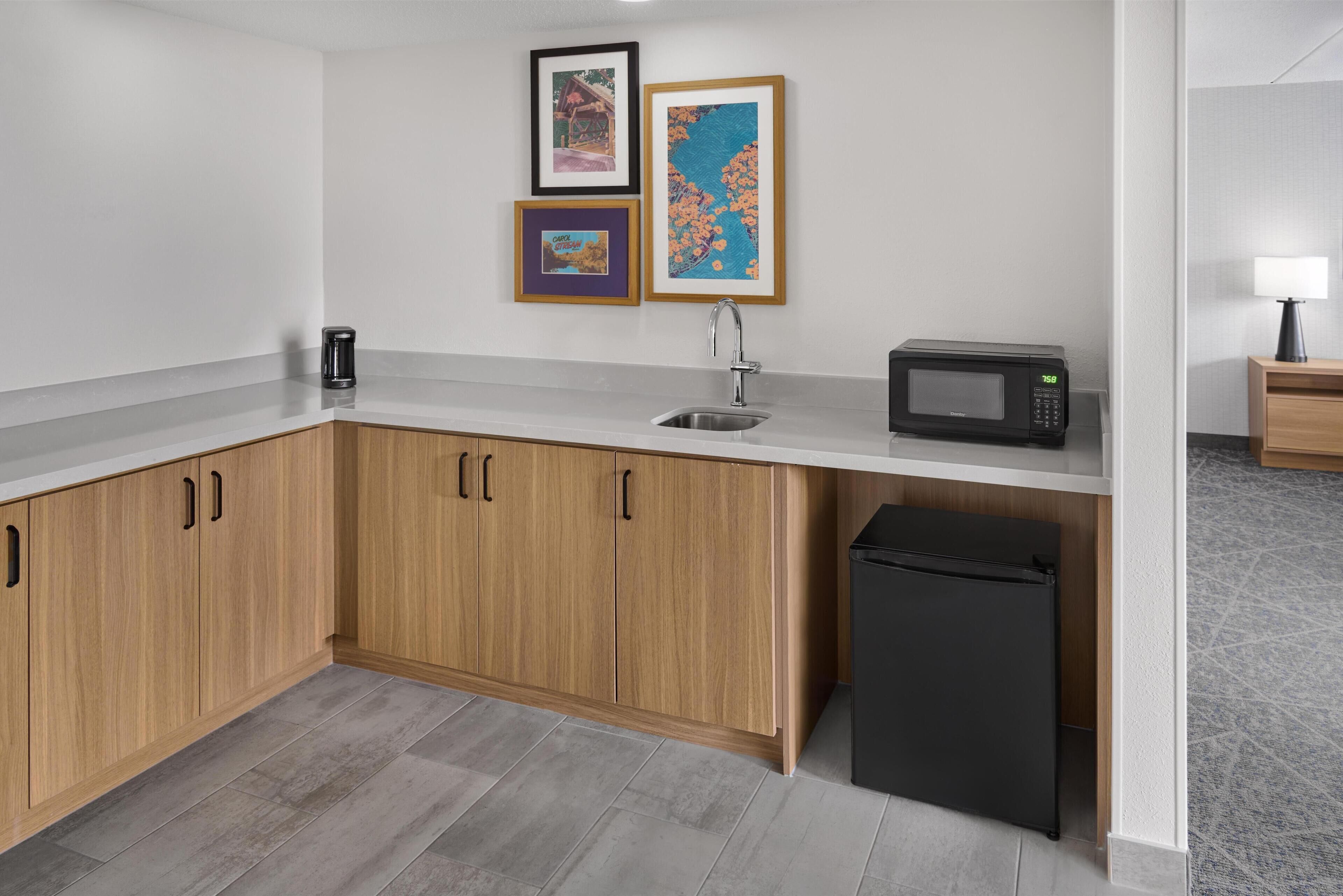 Une cuisine compacte avec des meubles en bois, un évier en acier inoxydable, un four à micro-ondes et un réfrigérateur noir. Des tableaux encadrés ornent le mur.