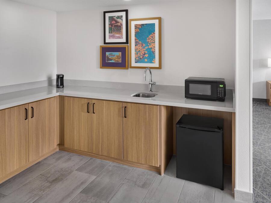 Eine kompakte Küche mit Holzausstattung, einem Edelstahlspülbecken, einer Mikrowelle und einem schwarzen Kühlschrank. An der Wand hängen gerahmte Bilder.