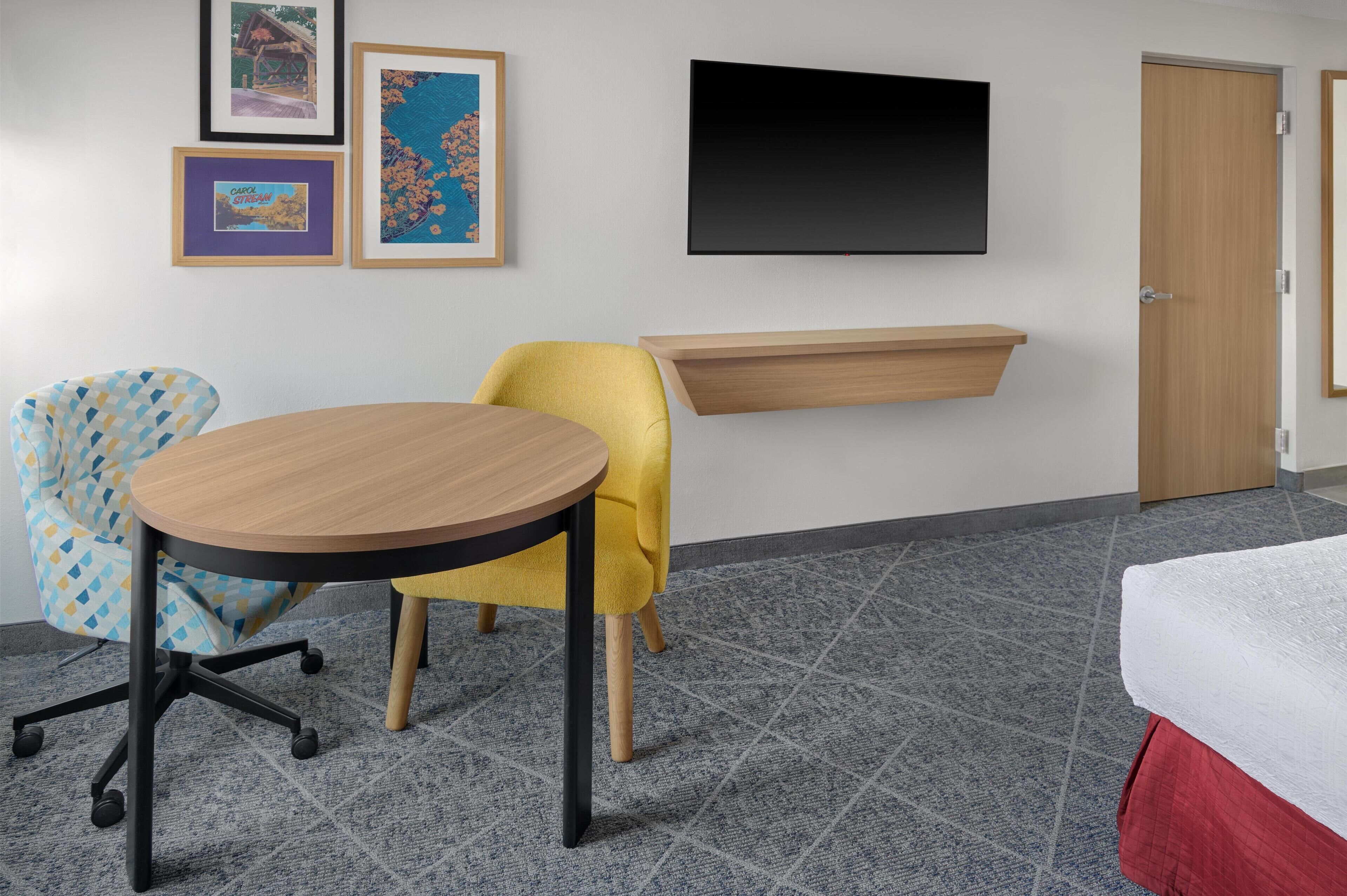 Una habitación con una mesa redonda, dos sillas, una cama, un televisor y cuadros enmarcados en la pared.