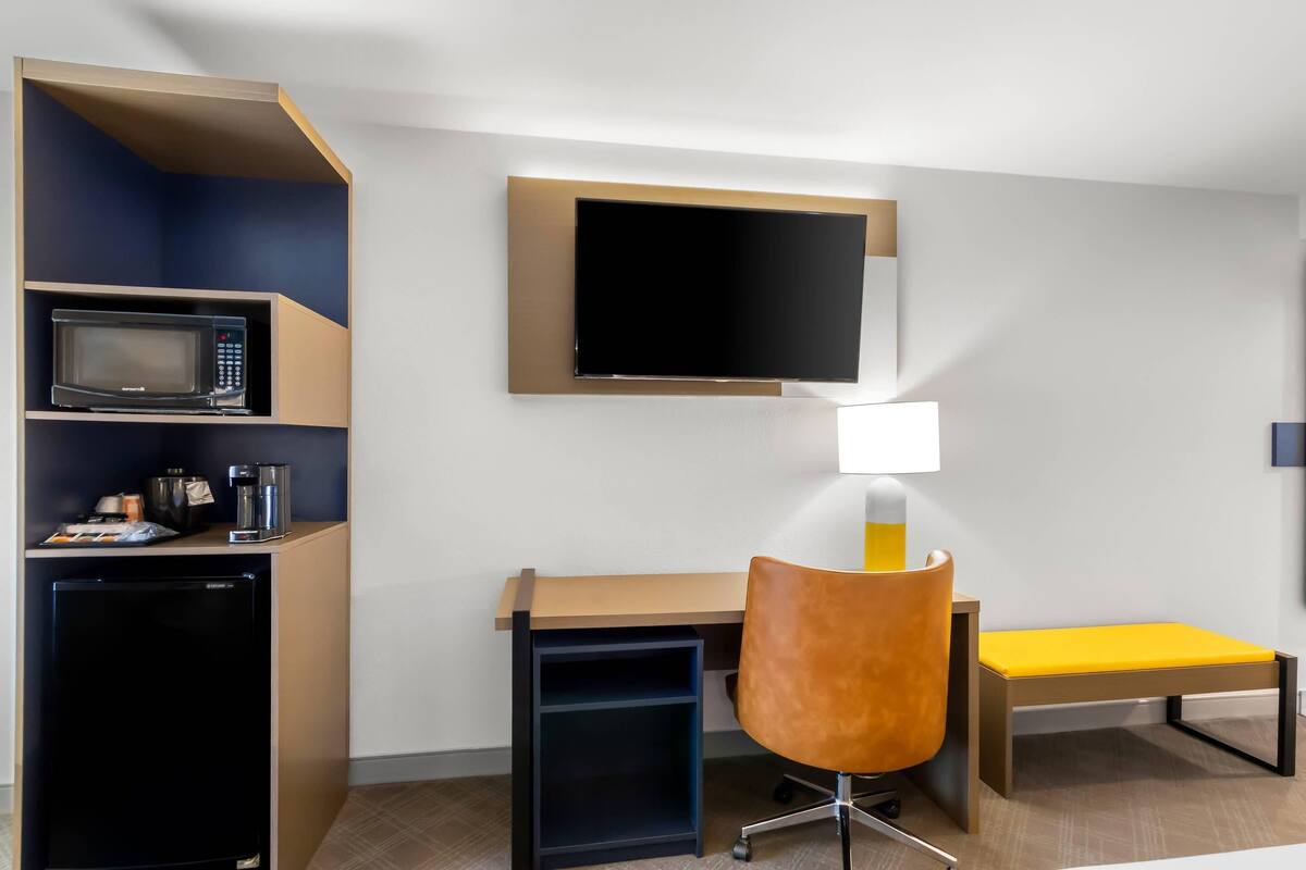 A hotel room with a desk, a chair, a TV mounted on the wall, a microwave, a coffee maker, and a small fridge.
