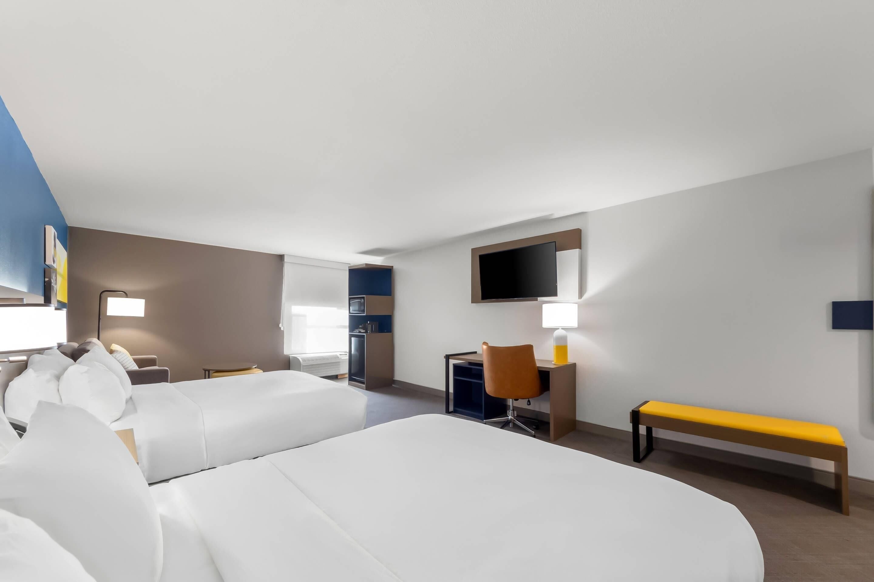 A hotel room with two beds, a desk, a chair, a TV, and a bench.