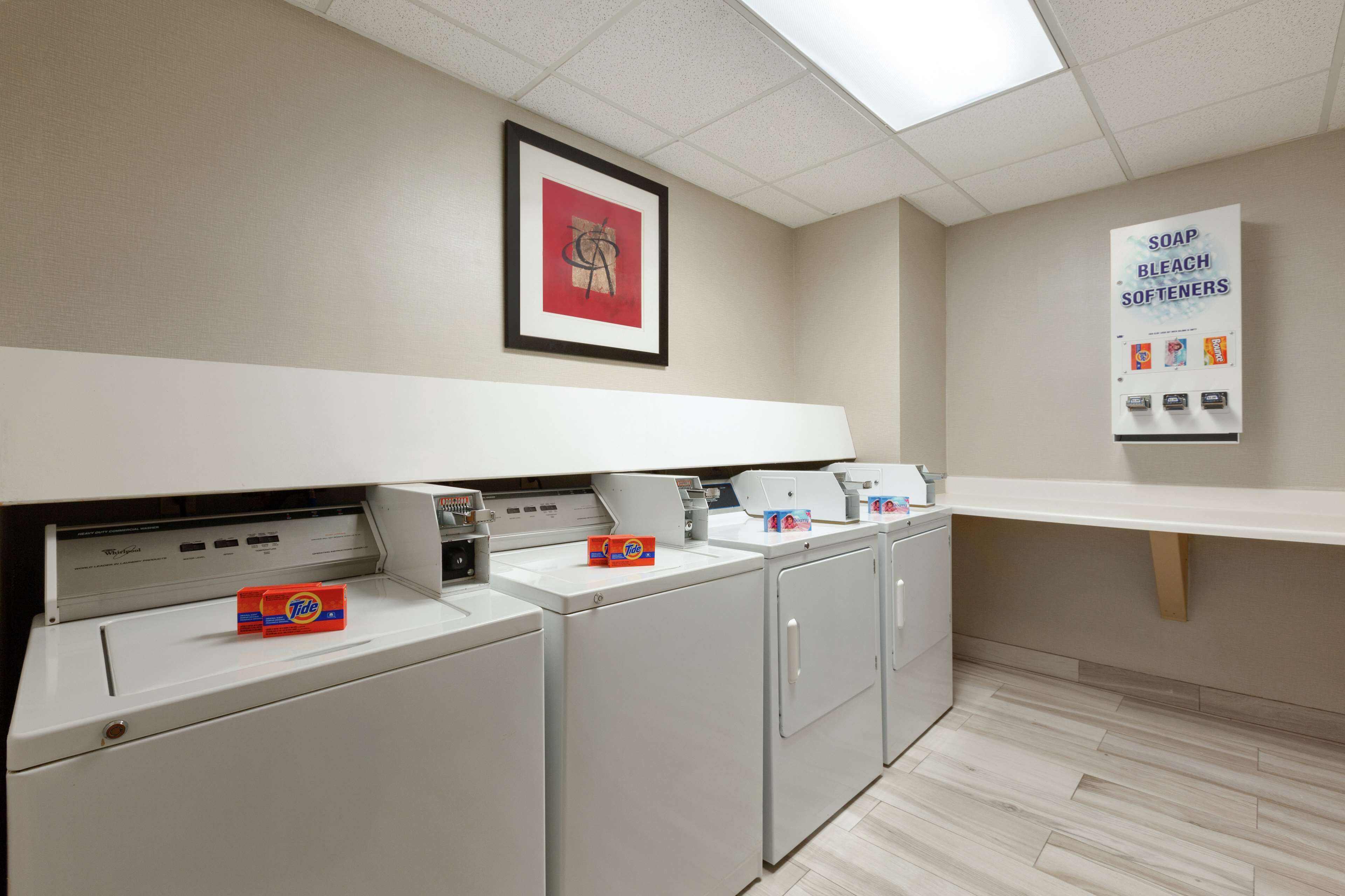 A laundry room with washers and dryers, a framed artwork on the wall, and a sign for soap and bleach softeners.