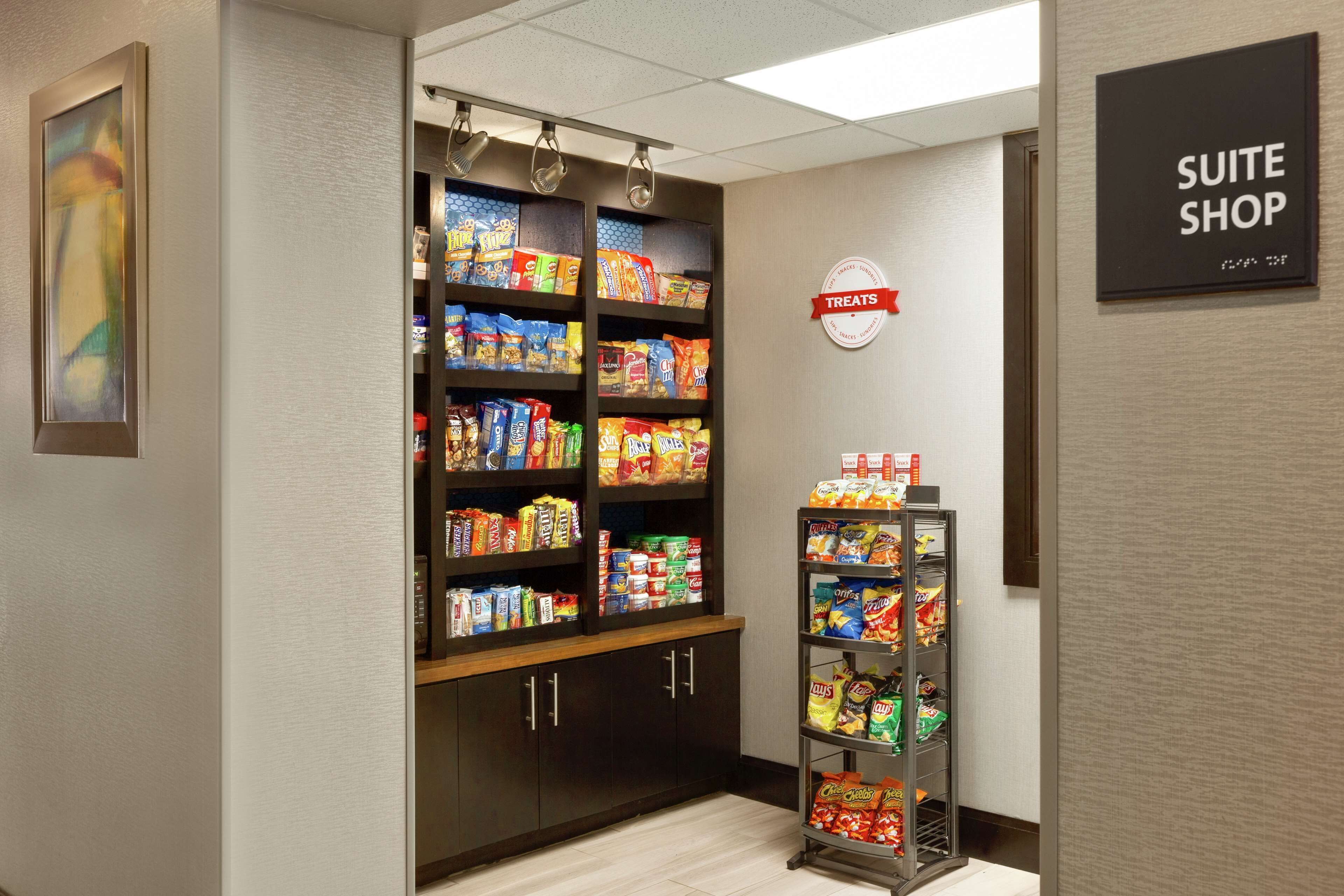 En hotellkorridor med ett litet köksutrymme, där hyllor är fyllda med snacks, och en skylt som visar vägen till hotellets butik.