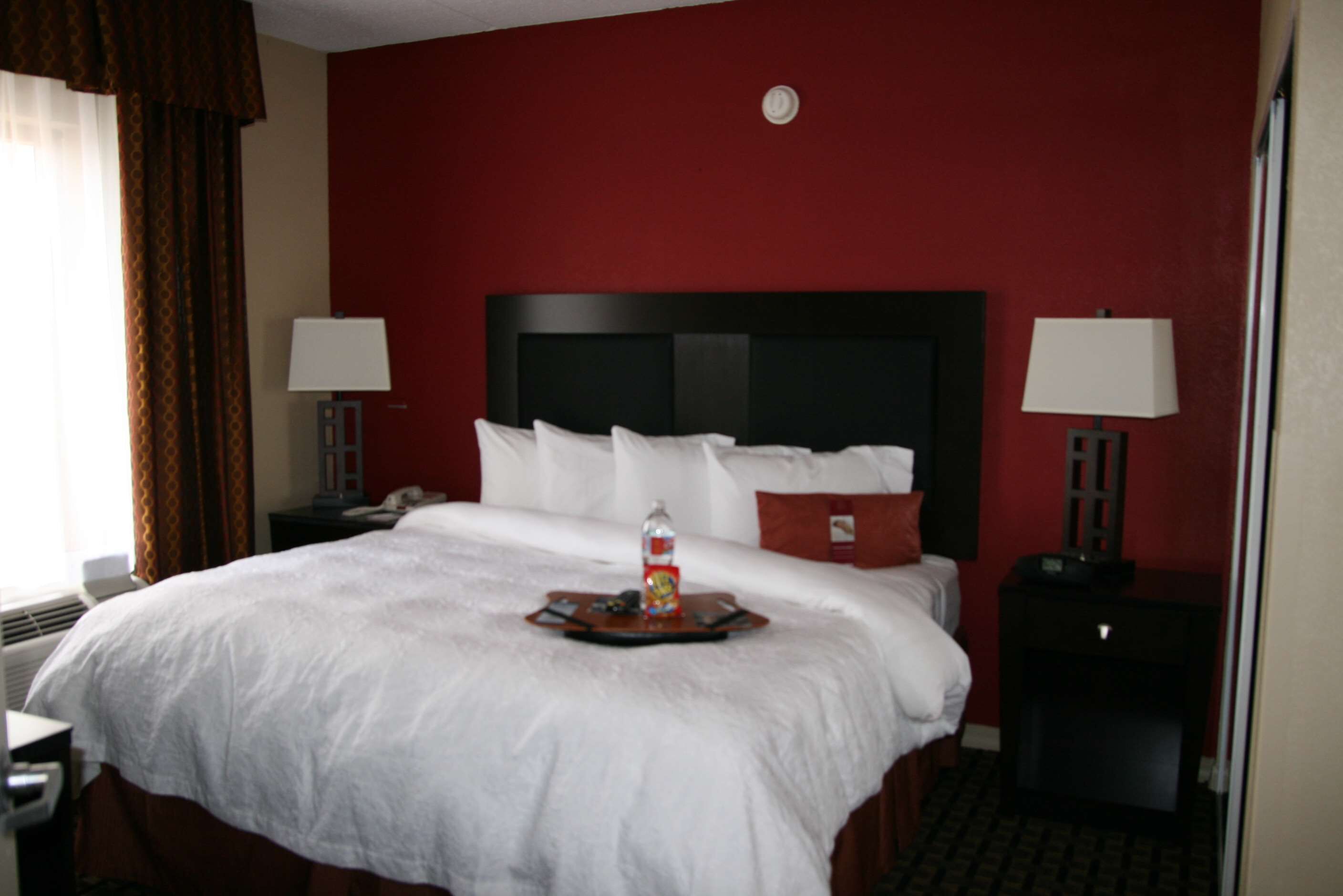 A hotel room with a large bed, two bedside tables with lamps, a red accent wall, and a tray with a bottle and snacks on the bed.