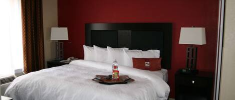 A hotel room with a large bed, two bedside tables with lamps, a red accent wall, and a tray with a bottle and snacks on the bed.