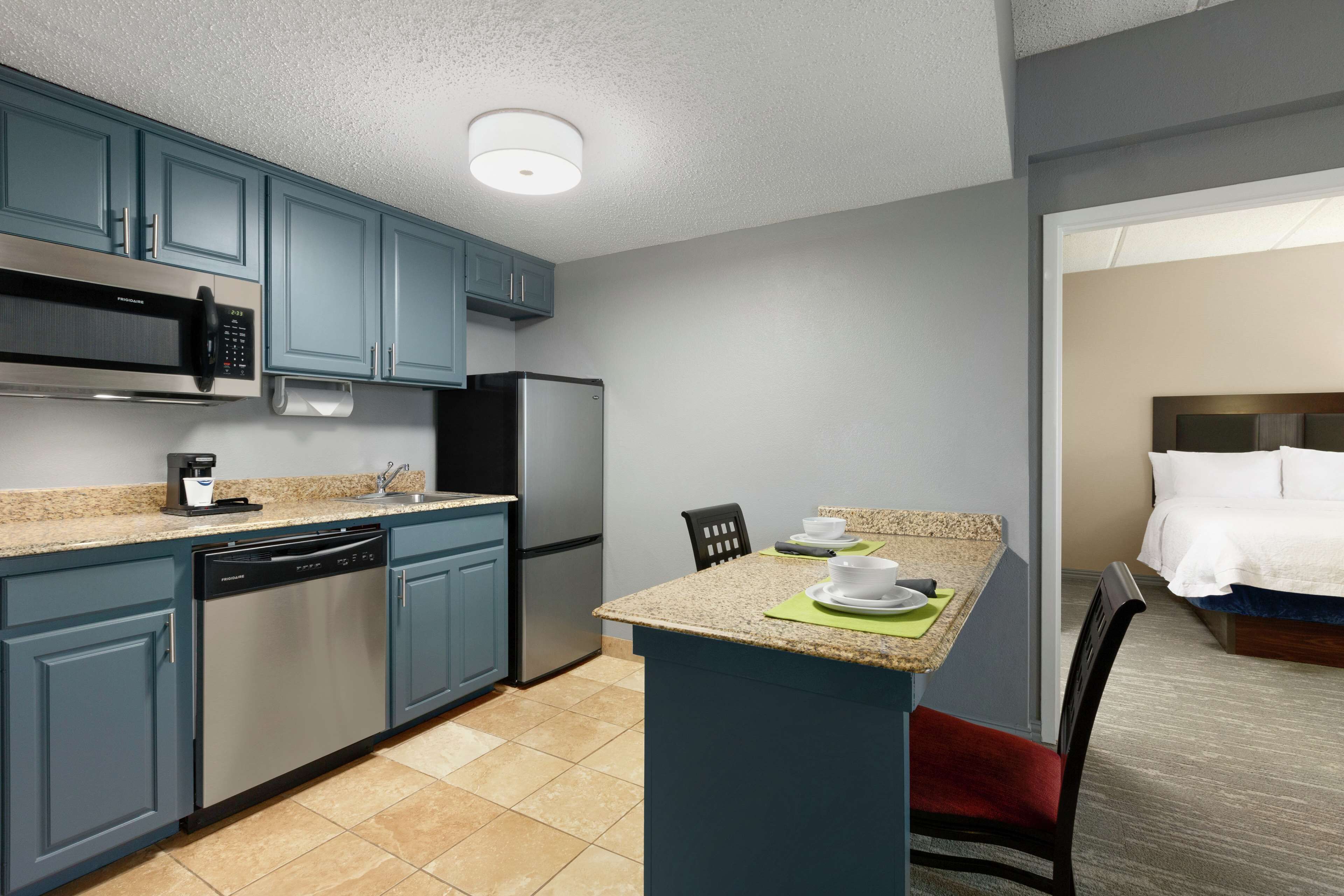 A compact kitchen with blue cabinets, a microwave, dishwasher, and a refrigerator. There is a small island with a sink and a chair.