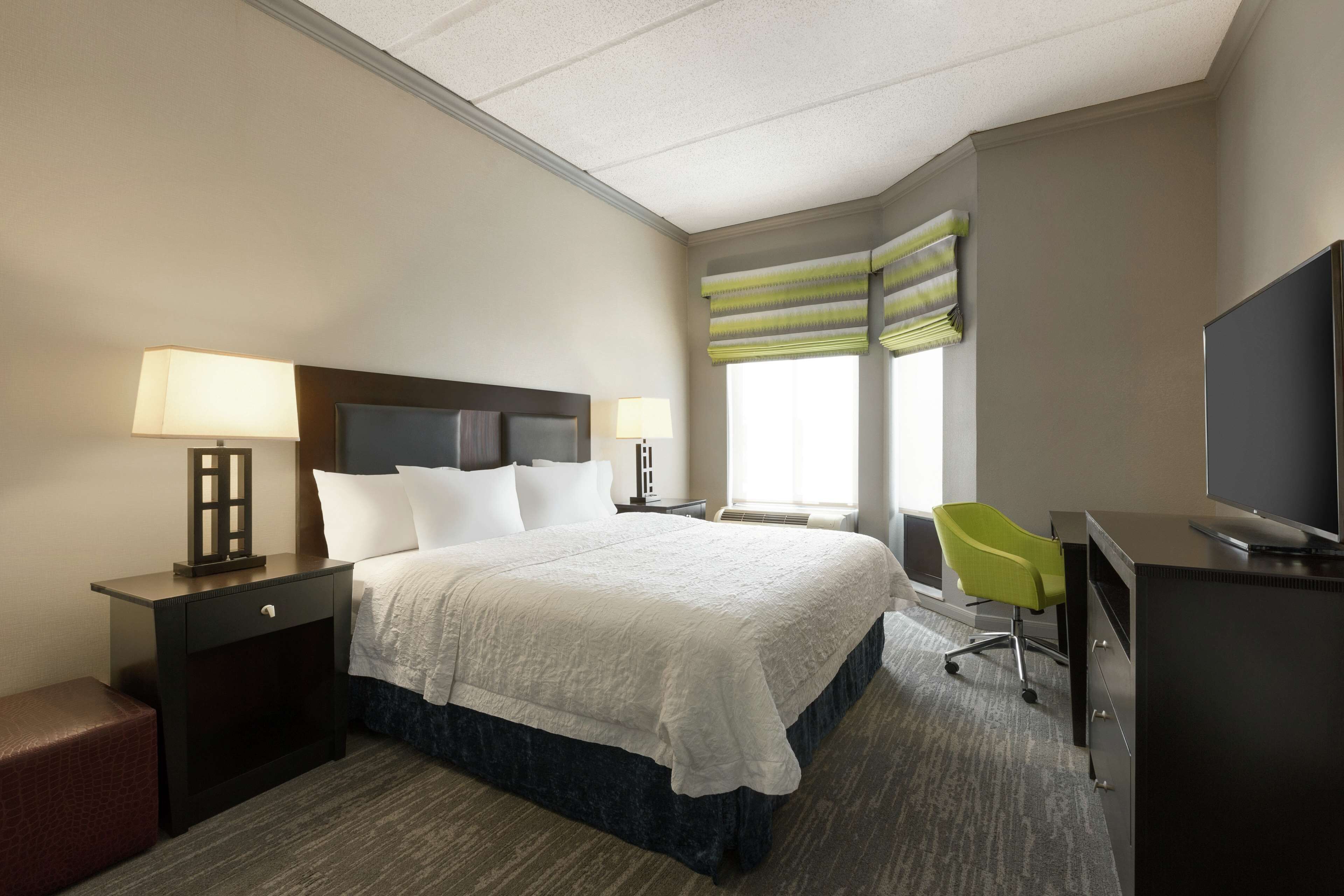 Habitación de hotel con cama, mesitas de noche, escritorio con silla, televisor y ventana con persianas.