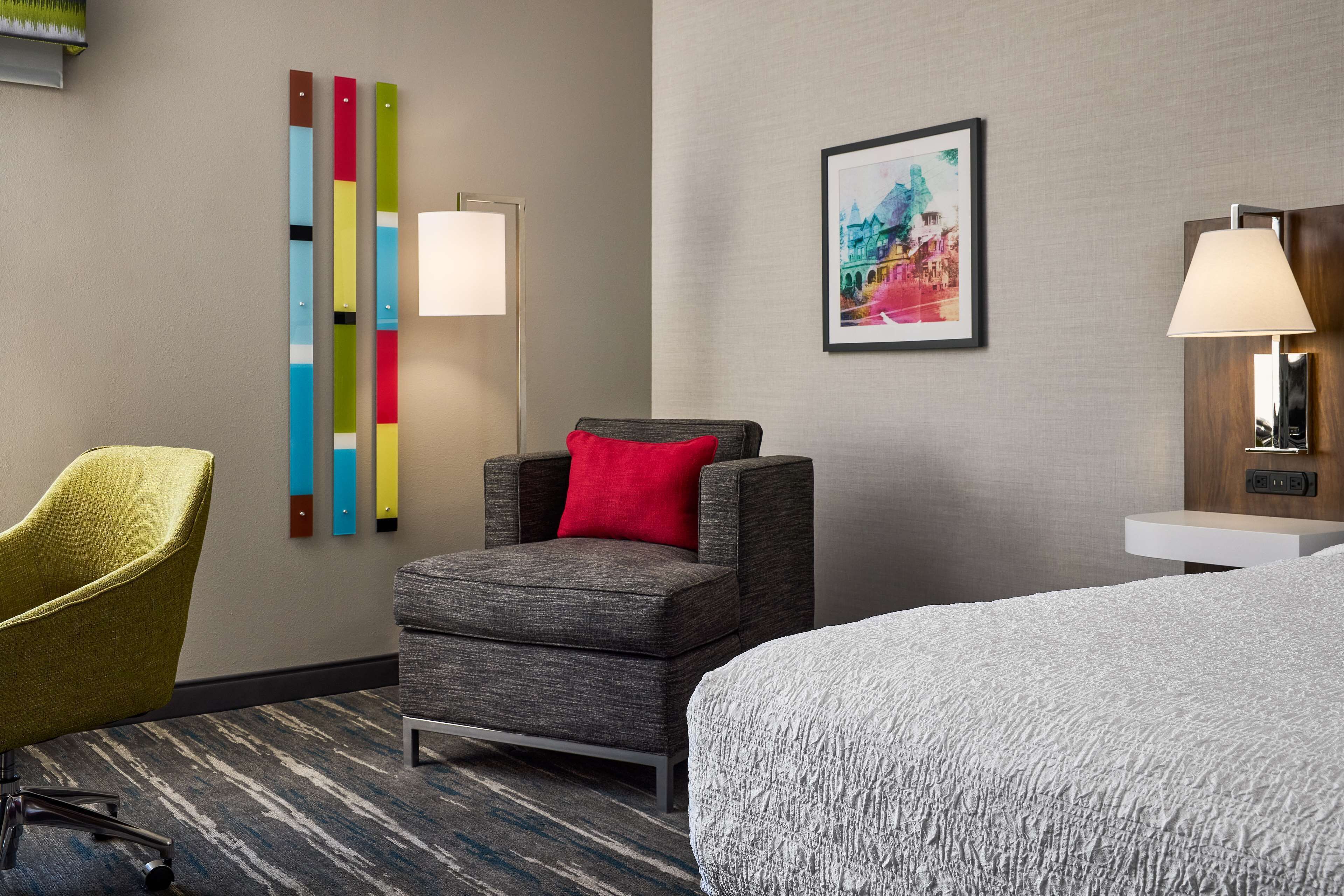 Ein Hotelzimmer mit grauem Sessel, einem gelben Stuhl, einem Bett mit strukturierter weißer Bettdecke, einer Wandlampe, einem gerahmten Bild und einem farbenfrohen abstrakten Kunstwerk.
