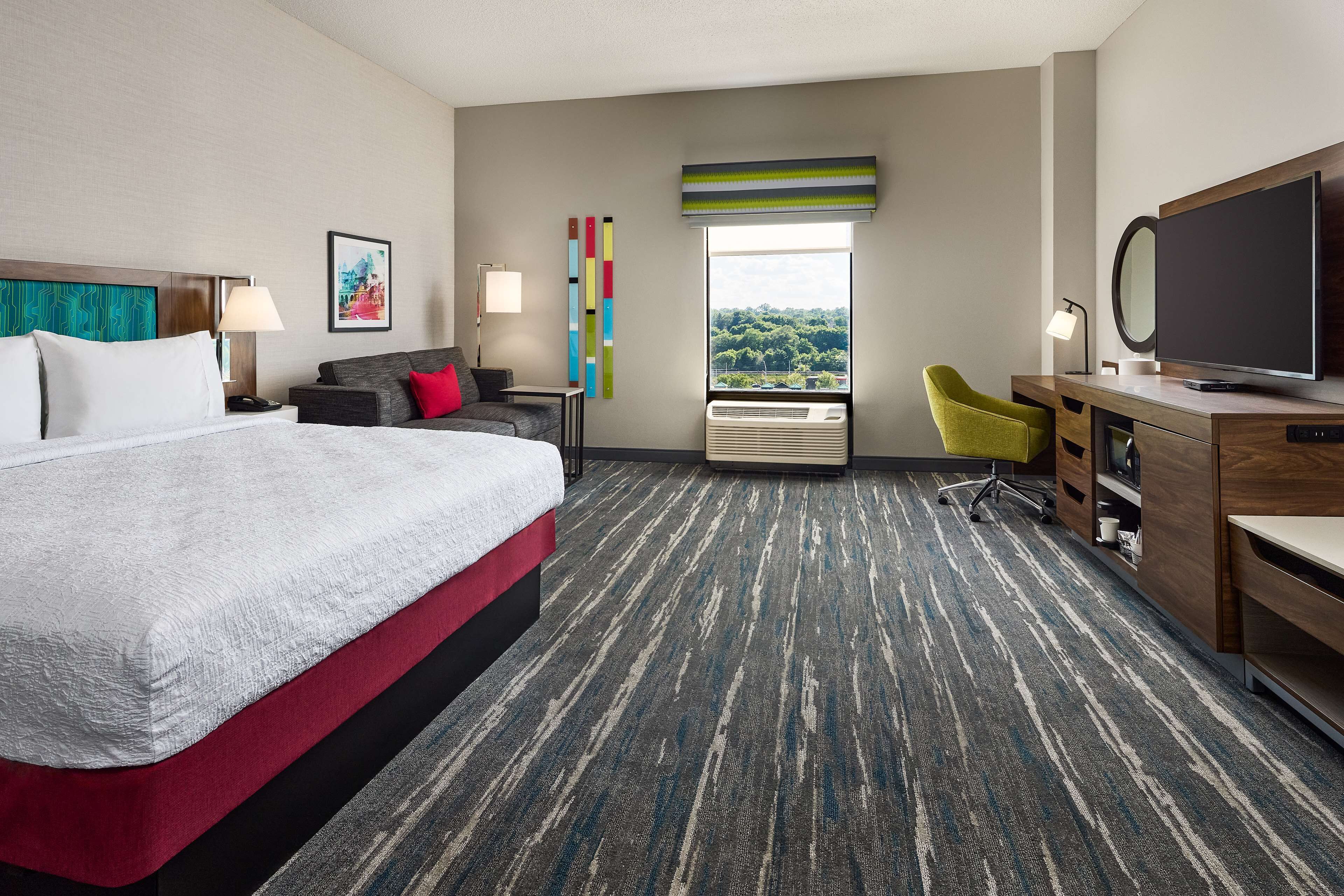 Habitación de hotel con cama, escritorio con televisor, silla, lámpara de escritorio, ventana con vistas y una pared decorada con un cuadro.