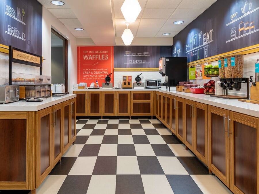 Een moderne keuken met een geruit vloertje, houten kastjes en een aanrecht waarop diverse apparaten en etenswaren staan.