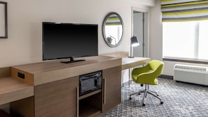 Une chambre d’hôtel avec un bureau en bois, un téléviseur à écran plat, un fauteuil vert, un miroir rond et une fenêtre avec des rideaux à rayures.