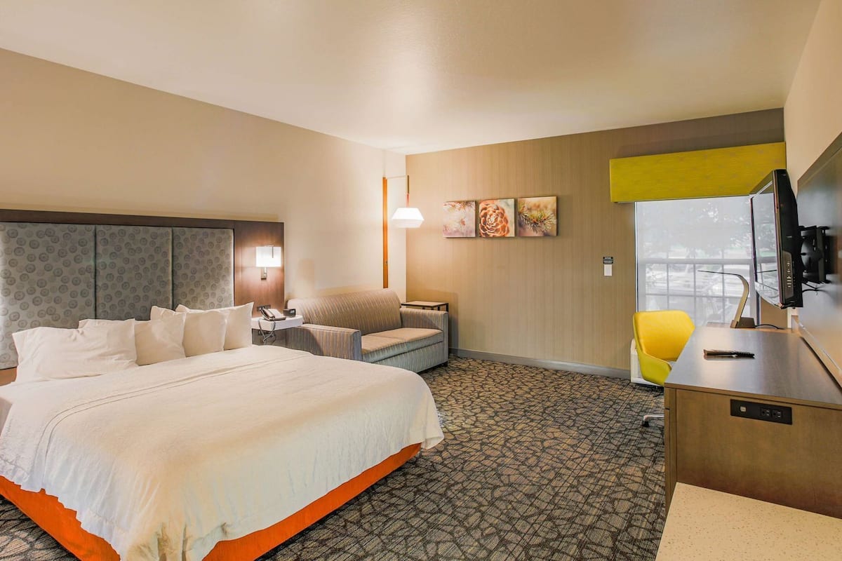 A hotel room with a large bed, a desk with a computer, a chair, and artwork on the wall.