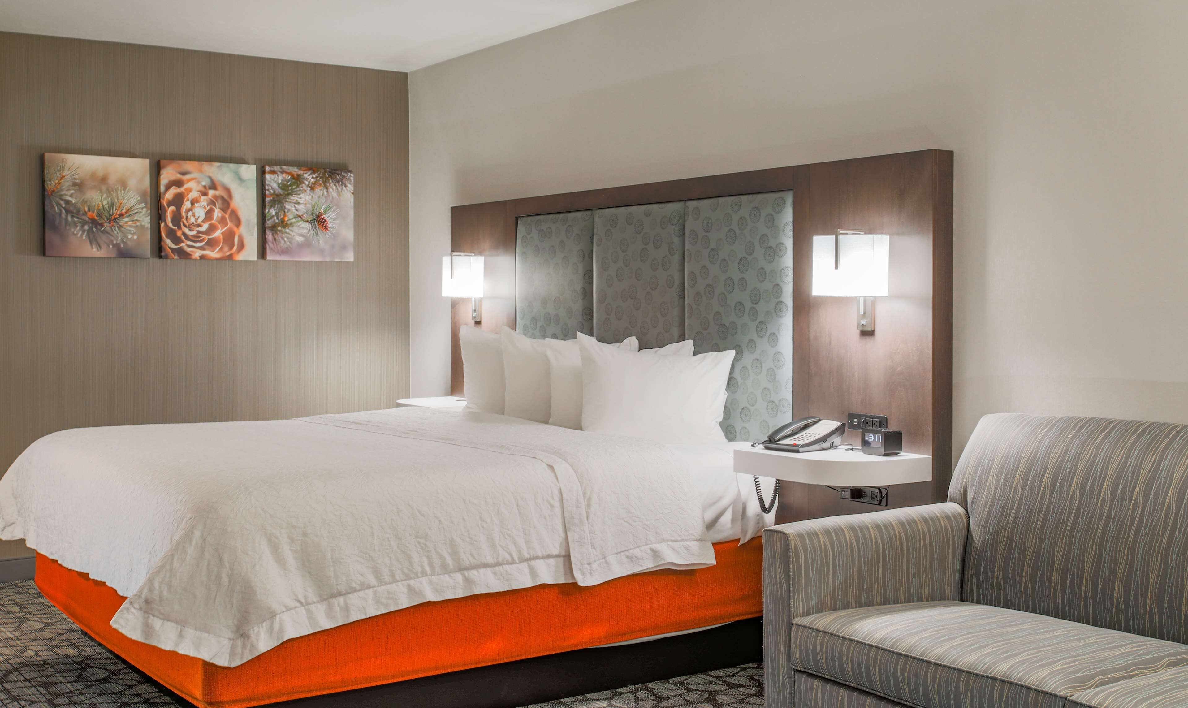 A hotel room with a bed, a chair, a bedside table with a phone and clock, and two wall-mounted lamps.