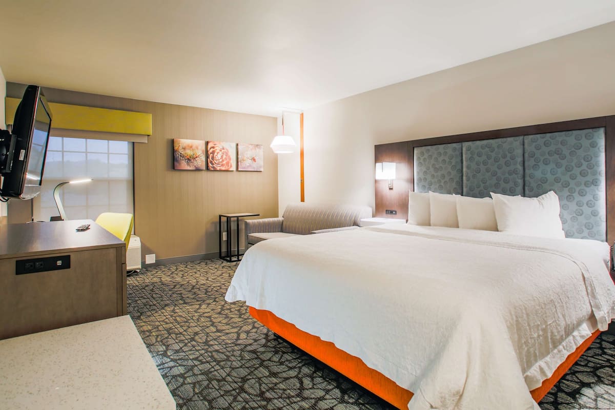 A hotel room with a large bed, a desk with a computer, a chair, a small table, and a wall-mounted TV.