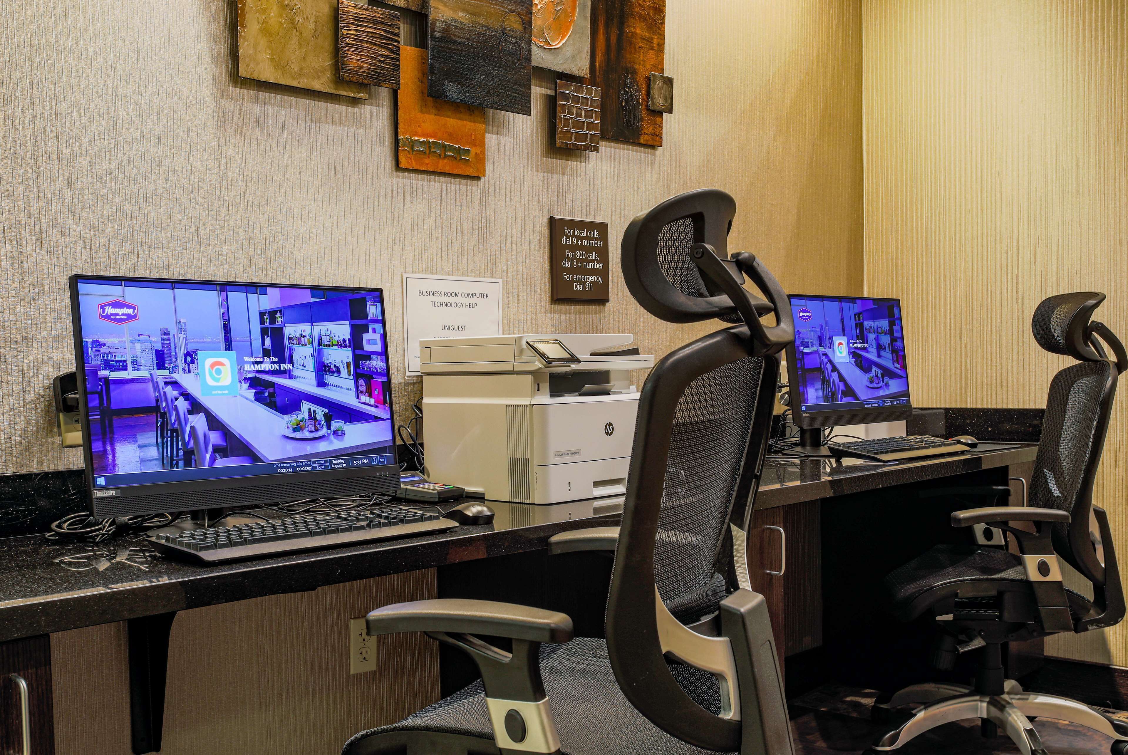 A hotel business room with two computer monitors, a printer, and office chairs.