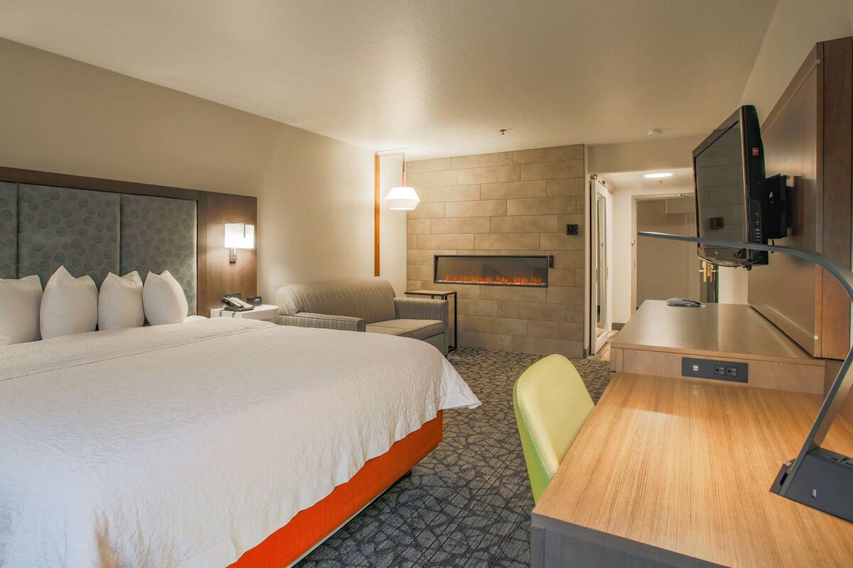 A hotel room with a large bed, a seating area with a chair and a small table, a flat-screen TV mounted on the wall, and a fireplace.
