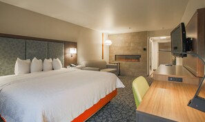A hotel room with a large bed, a seating area with a chair and a small table, a flat-screen TV mounted on the wall, and a fireplace.