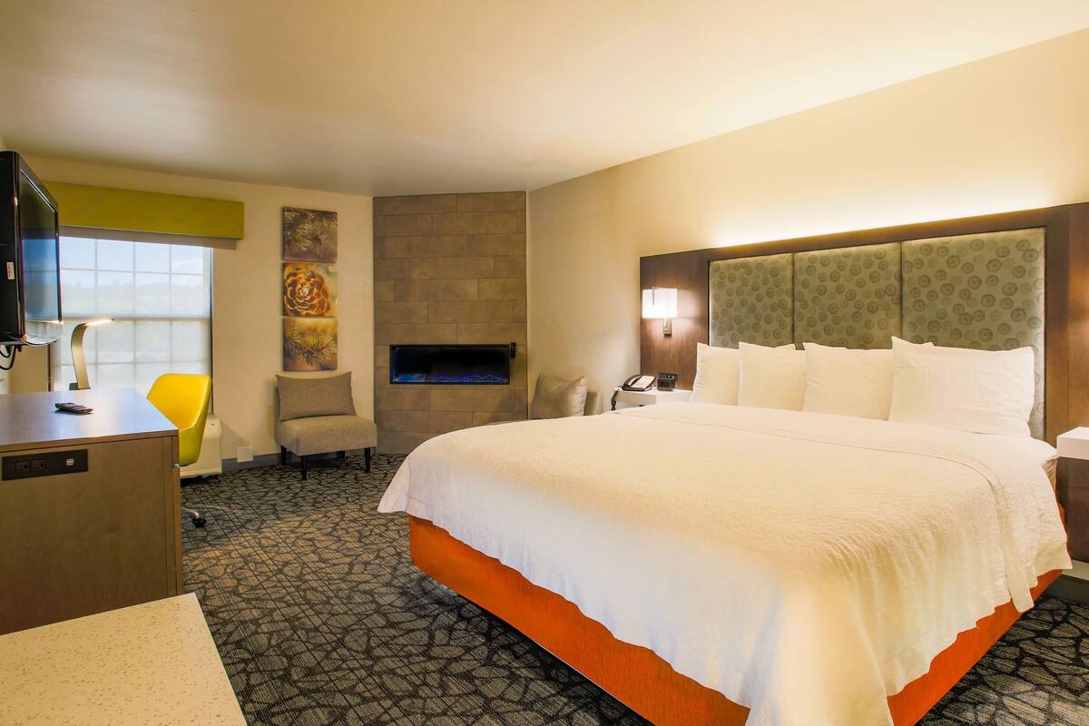 A hotel room with a large bed, a desk with a computer, a chair, a fireplace, and a television.