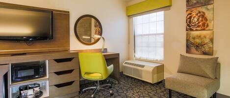 A hotel room with a desk, a television, a mirror, a window, and artwork on the walls.