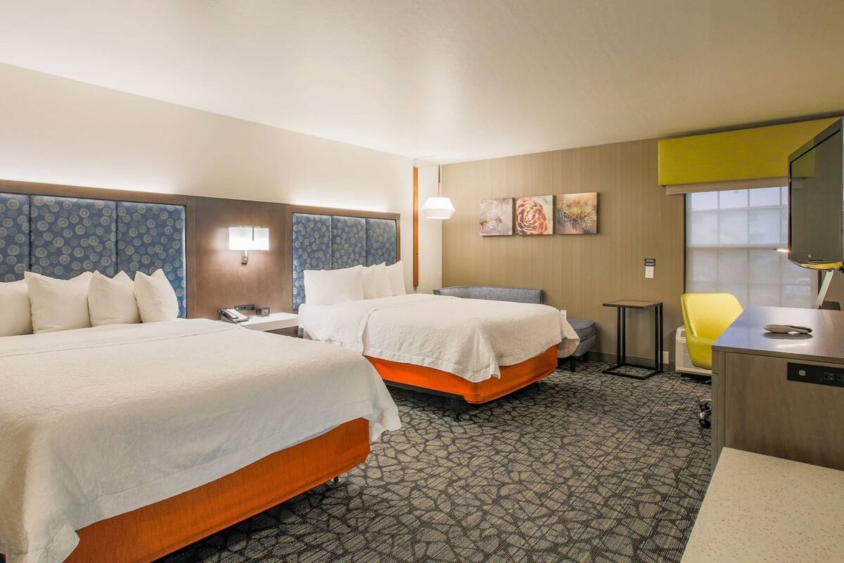A hotel room with two beds, a desk, a chair, a television, and artwork on the wall.