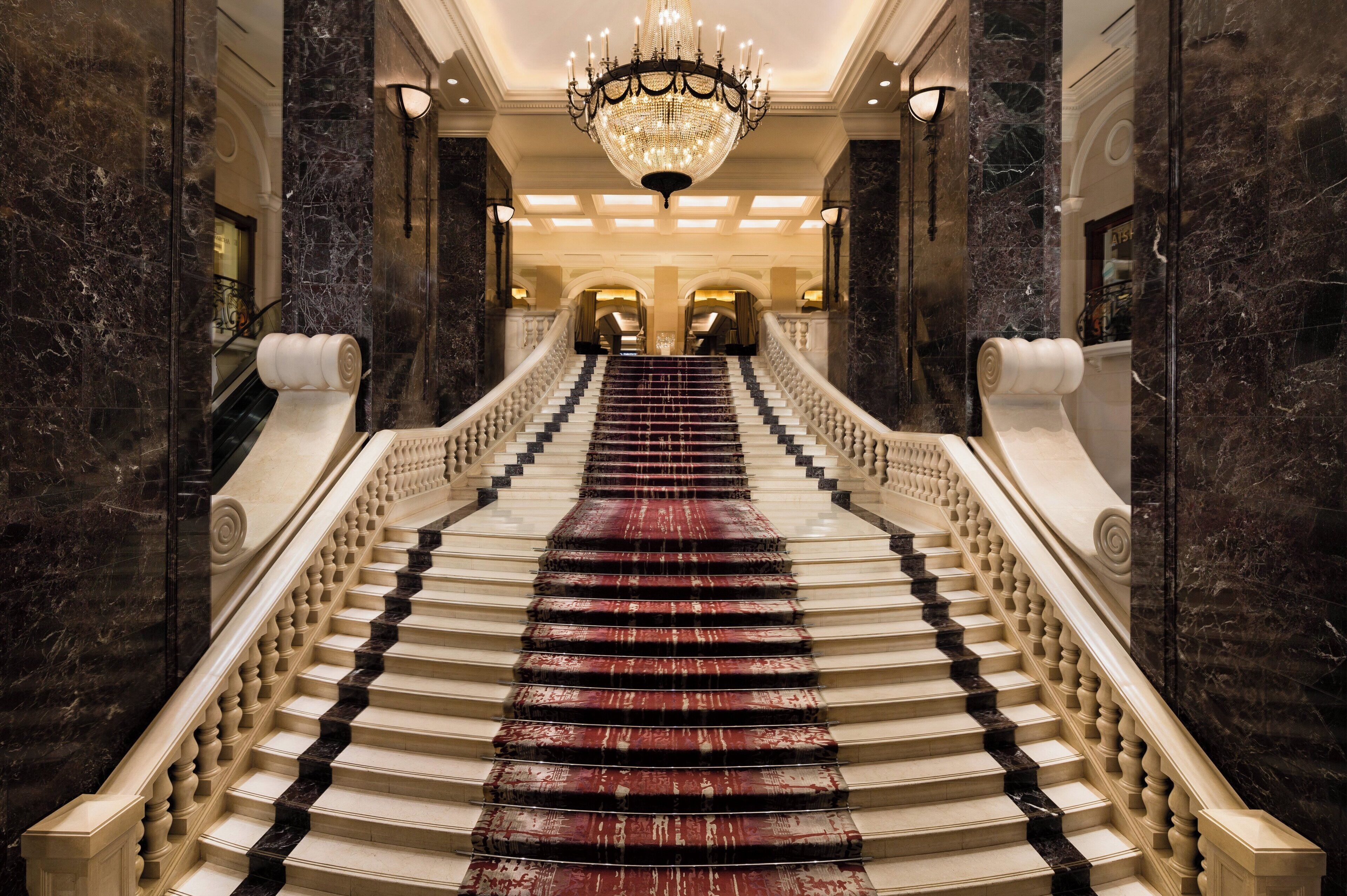 A grand staircase with marble steps and balustrades, leading to a well-lit area with a chandelier.